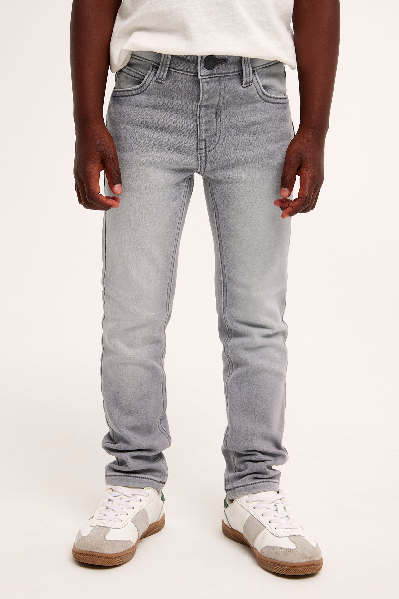 Framifrån: ett barn i gråa slim-fit joggerdenim jeans, en vit t-shirt och vita sneakers.