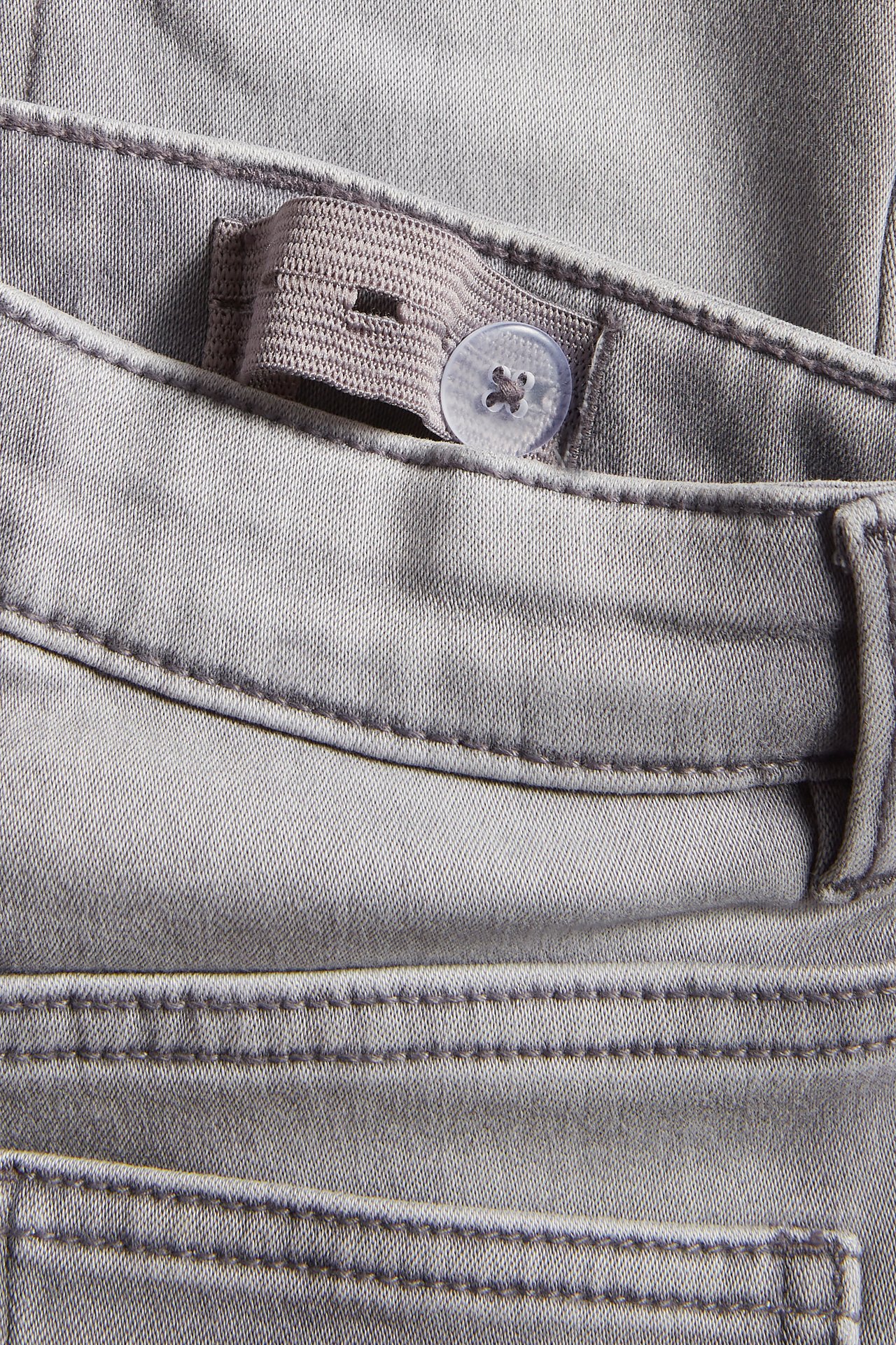 Närbild av gråa slim joggerdenim jeans, visar justerbar resårmidja med en genomskinlig knapp.