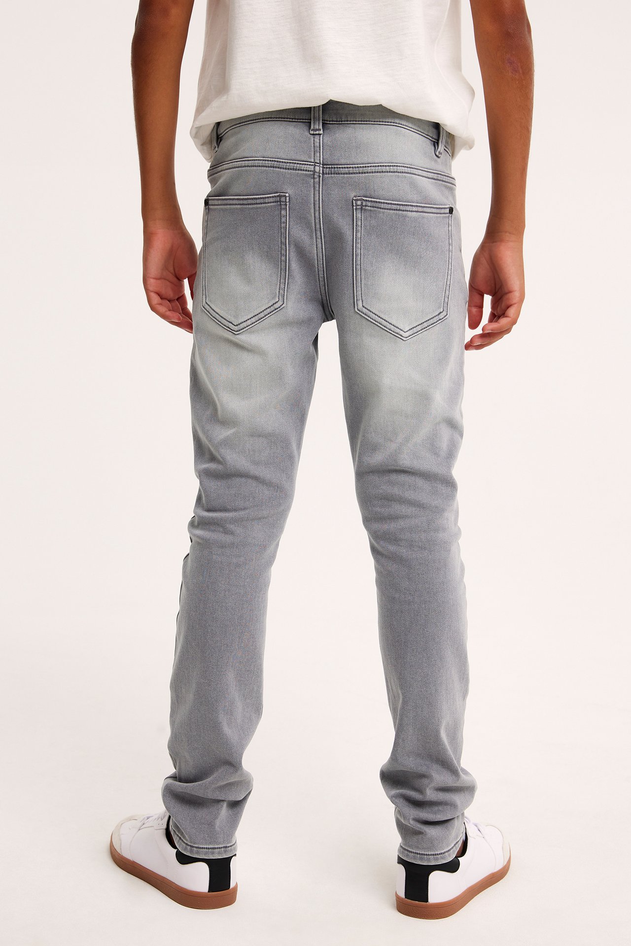 Bakifrån: ett barn i gråa slim joggerdenimjeans, en vit t-shirt och vita sneakers.