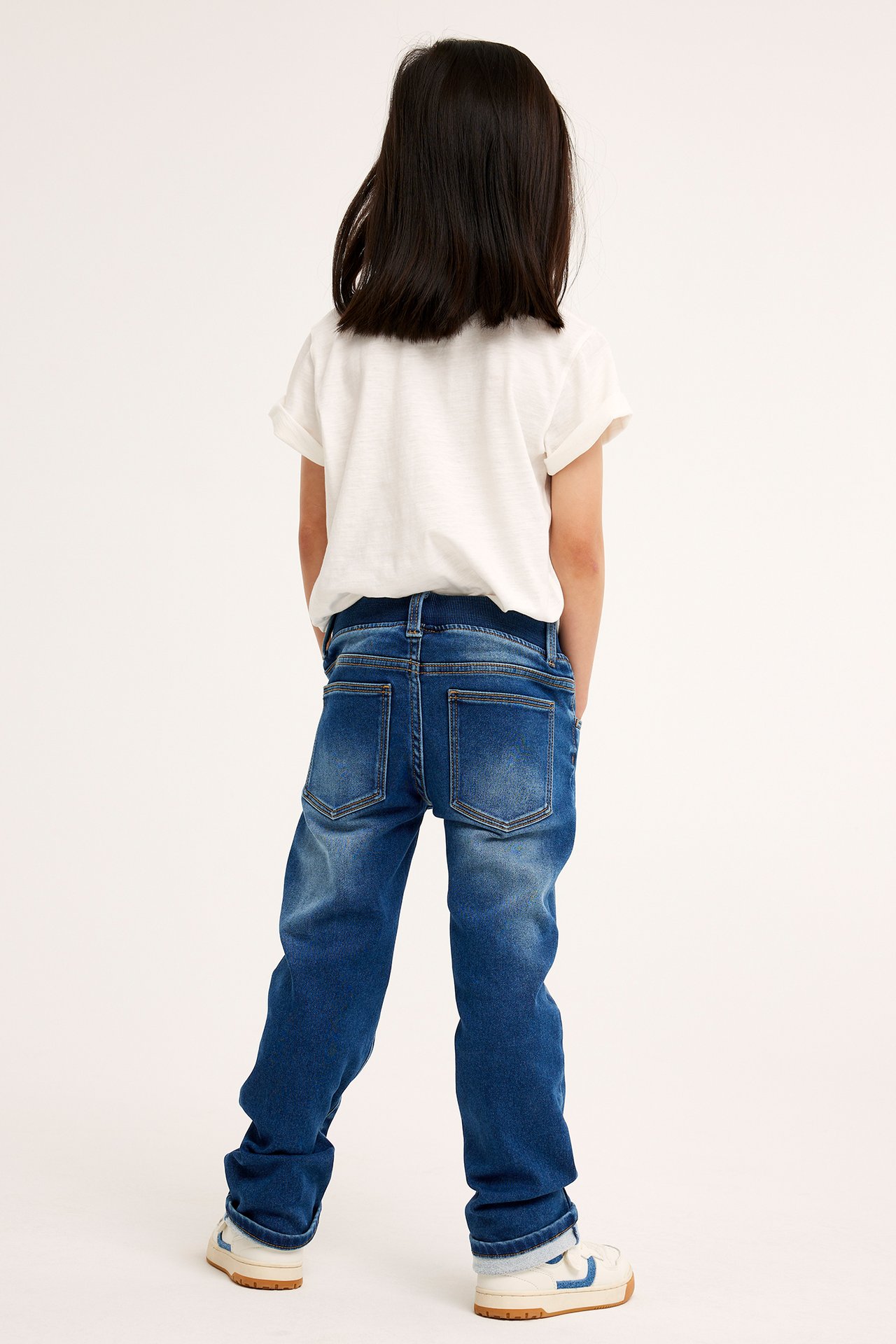 Bakifrån, ett barn i mörkblå raka joggerjeans, en vit t-shirt och vitblå sneakers.