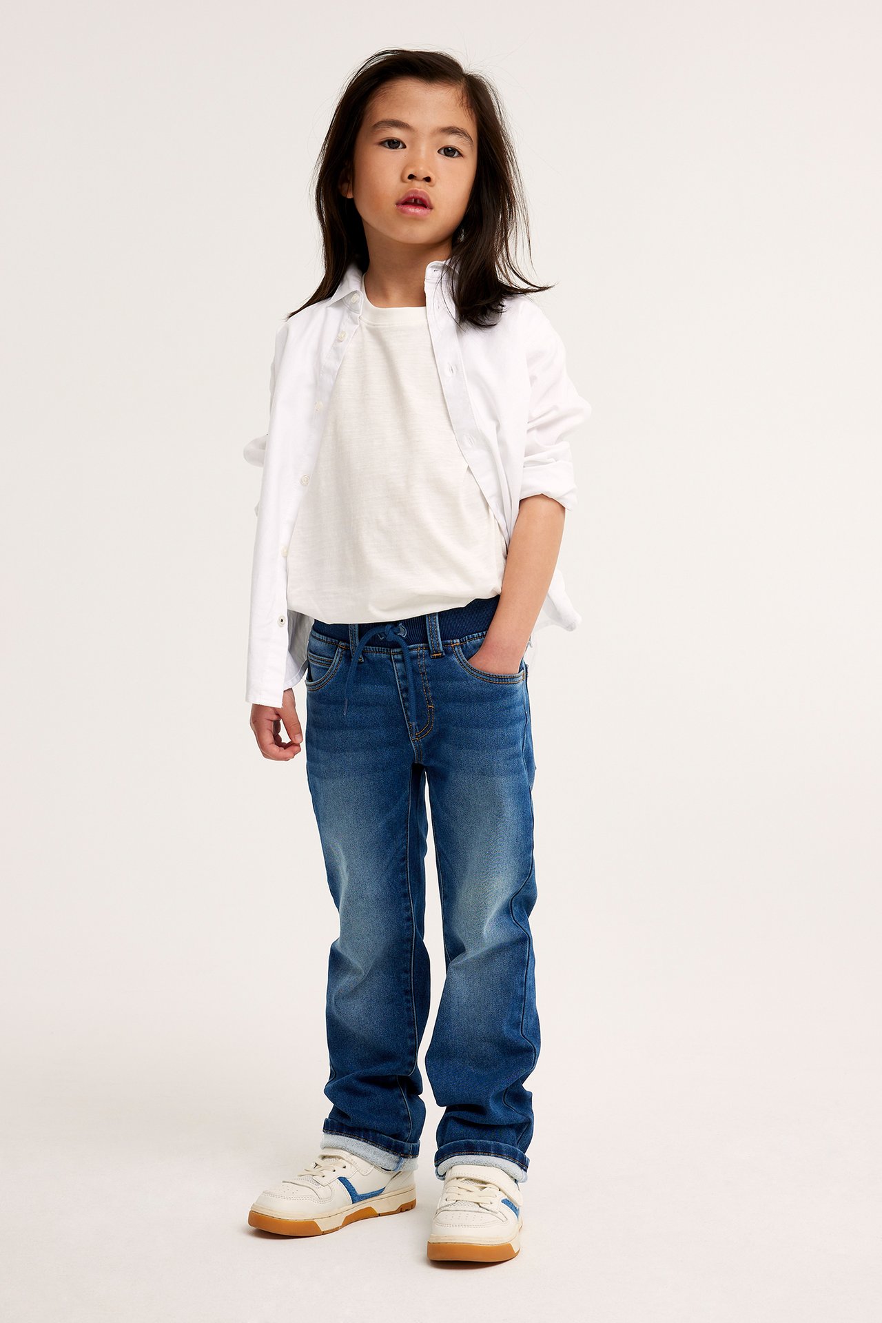 Frontvy av ett barn i mörkblå raka joggerjeans, ljusbeige t-shirt, öppen vit skjorta och vita sneakers.