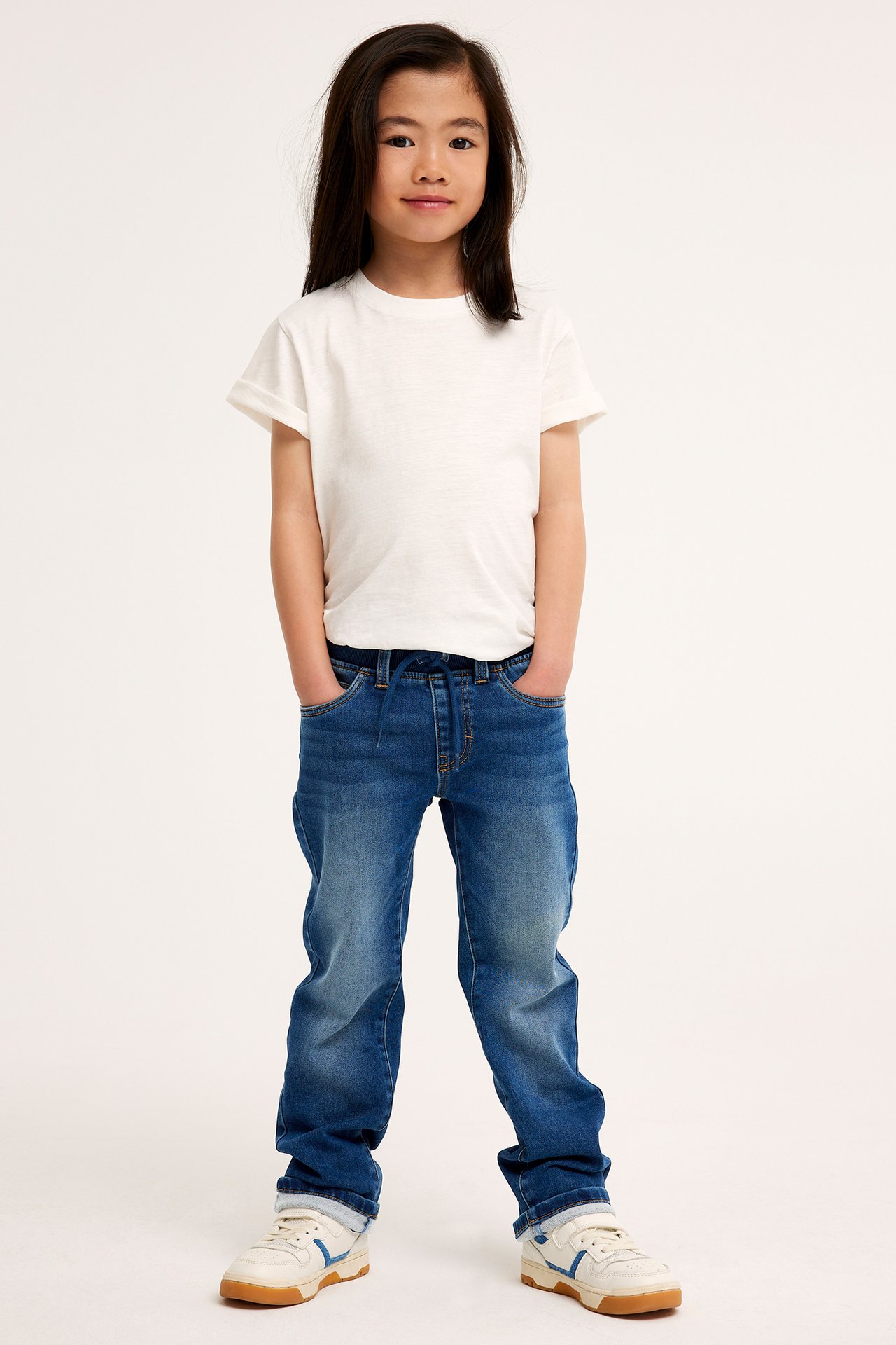 Frontvy av ett barn i mörkblå raka joggerjeans med dragsko, en vit t-shirt och vita sneakers.