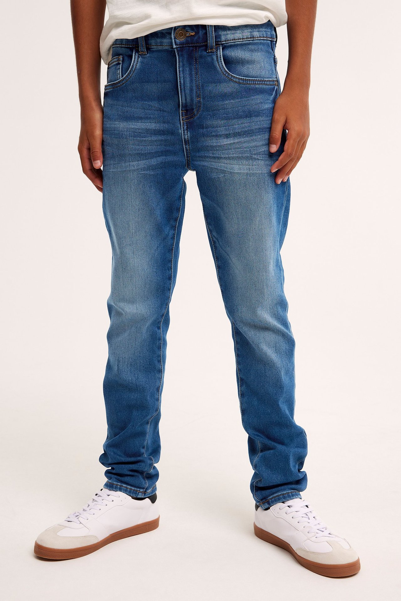 Edestäpäin, lapsi yllään siniset relaxed jogger denimfarkut, valkoinen t-paita ja valkoiset tennarit.
