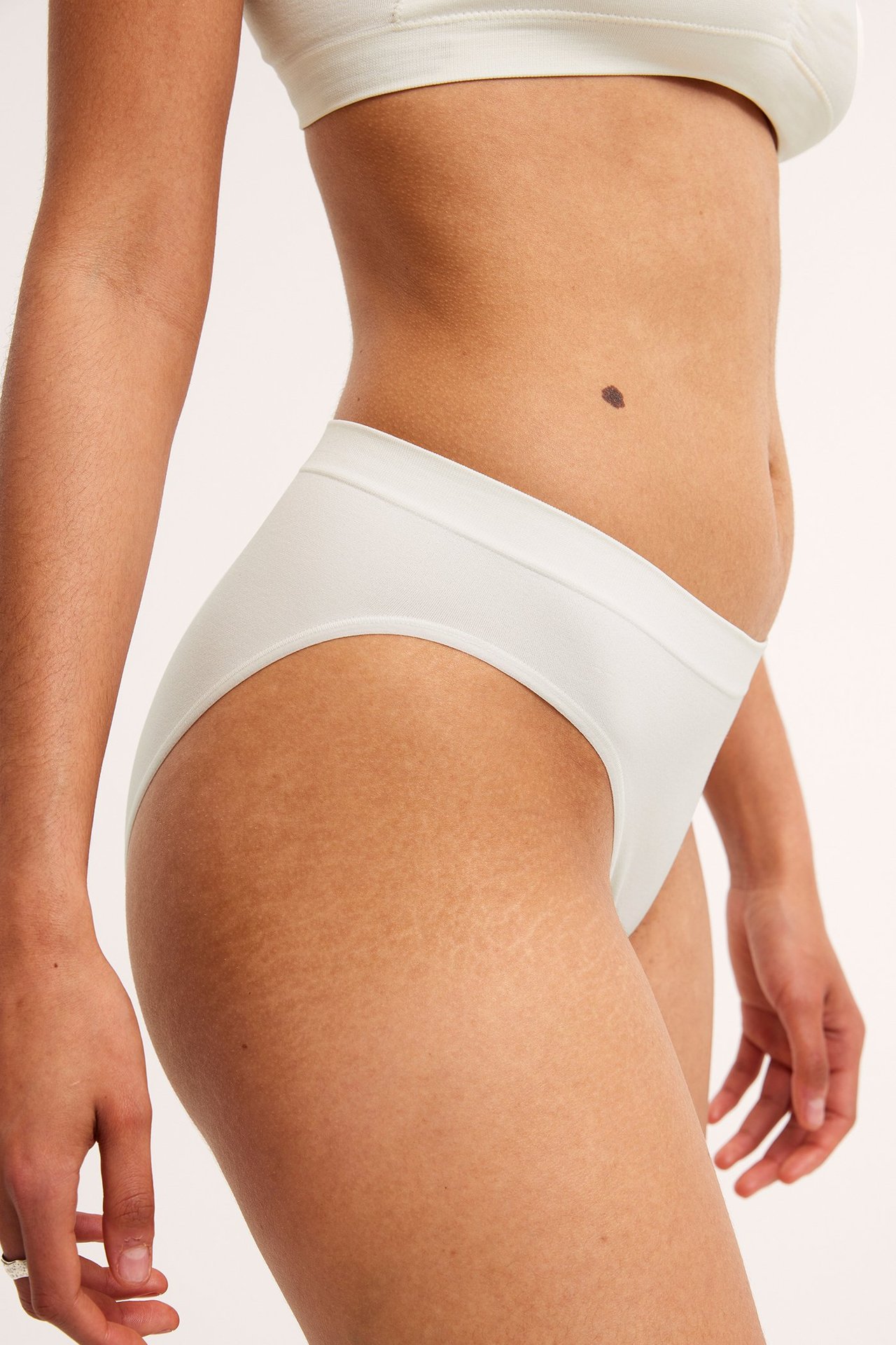 Sidebilde av en kvinne i hvite, myke truser med ribbet midje og matchende hvit bralette.
