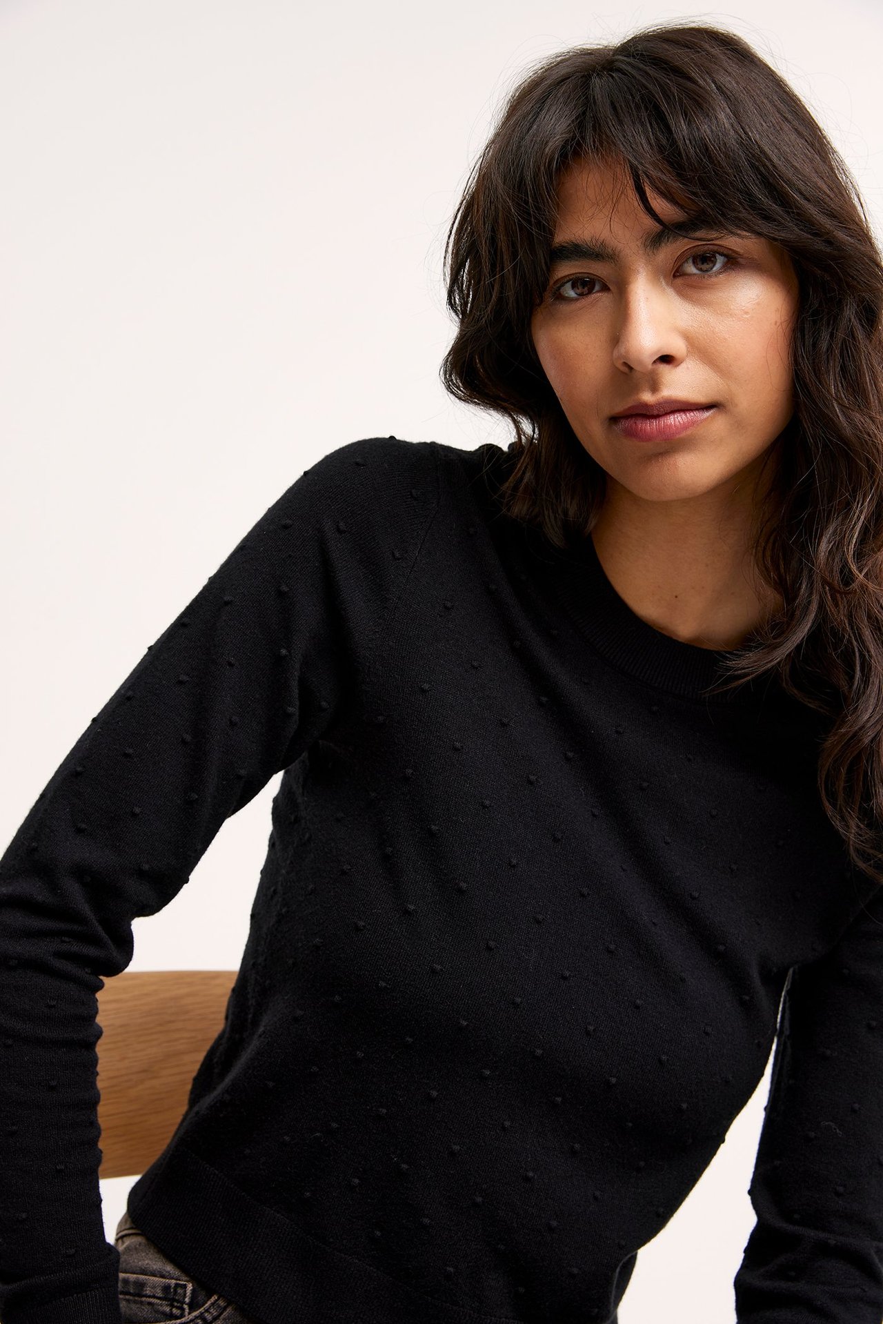 Lähikuva edestä naisesta yllään musta neulepusero hienolla pilkkukuviolla ja pitkillä hihoilla.