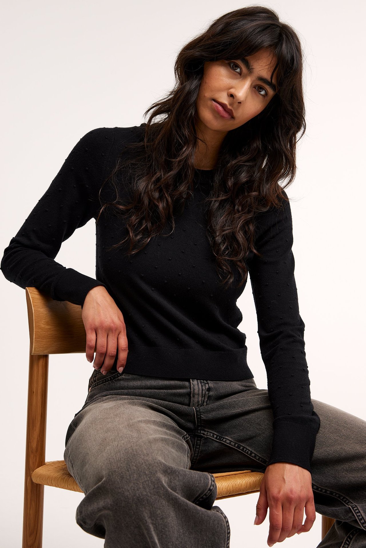 Naisen etunäkymä mustassa struktuurineulepuserossa ja löysissä harmaissa farkuissa.