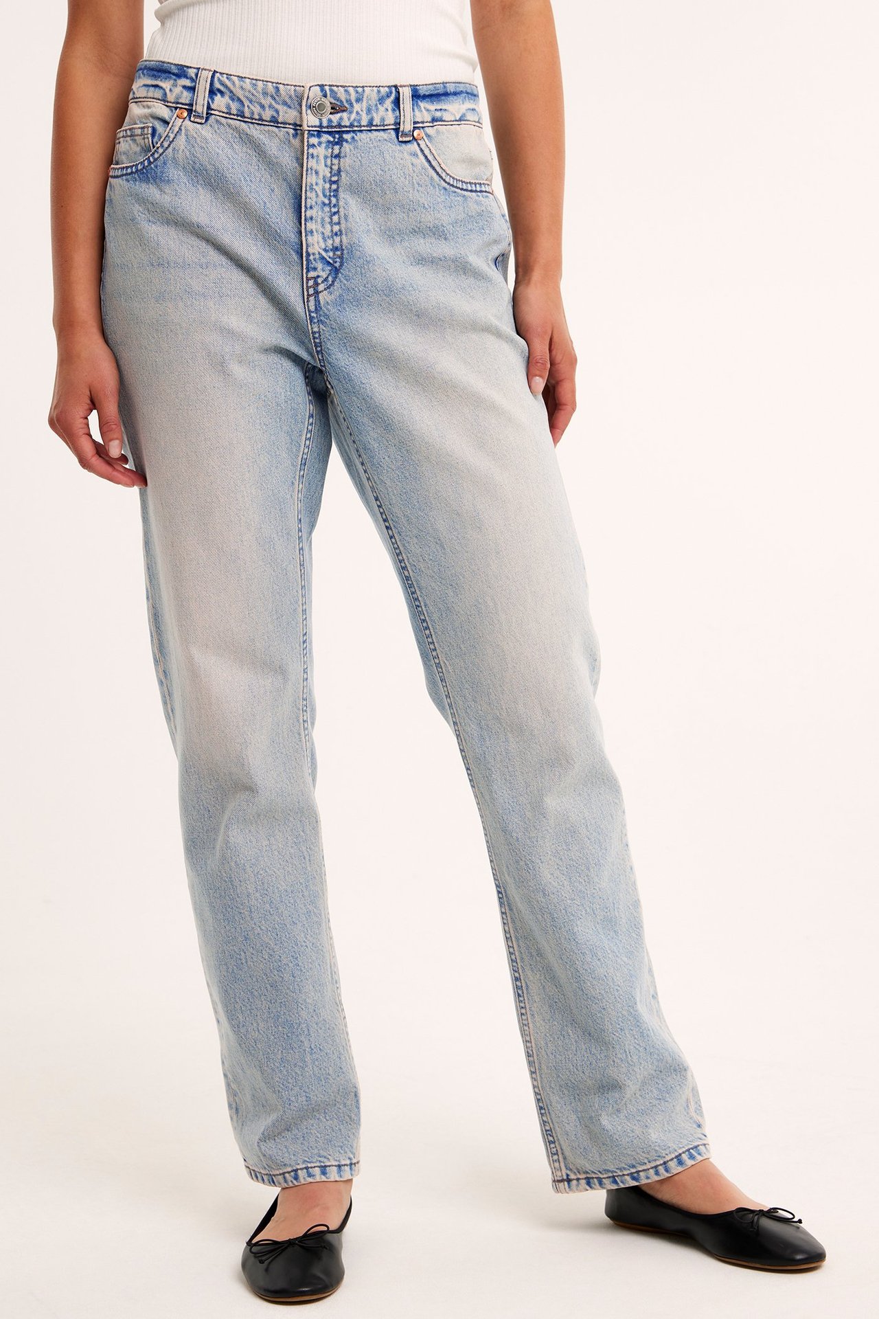 Frontvy av kvinna i ljusblå raka jeans med hög midja, vit ribbad topp och svarta ballerinaskor.