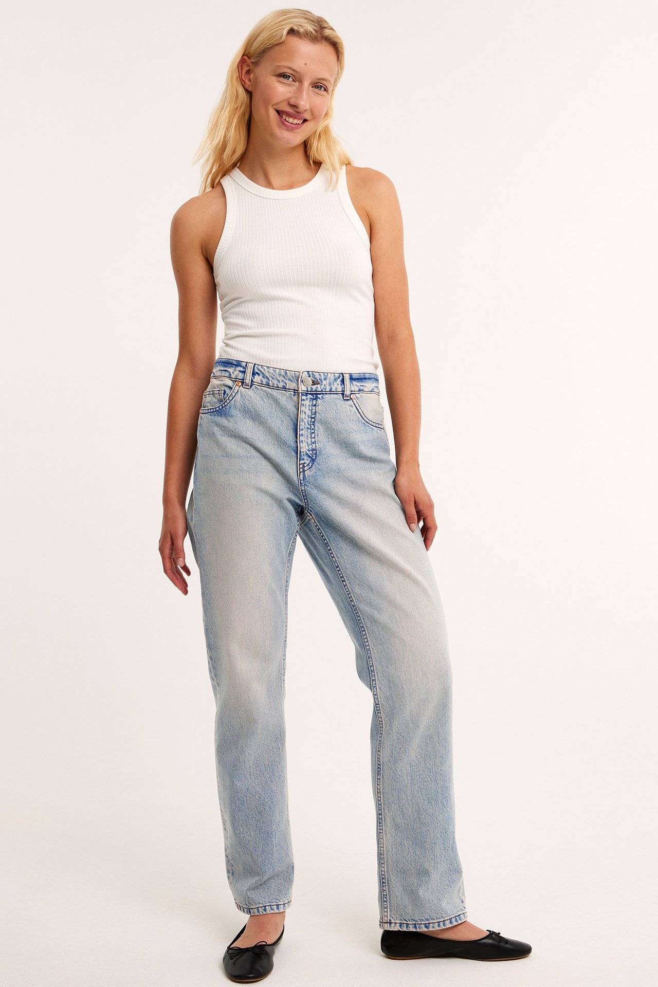 Framifrån: kvinna i ljusblå raka jeans med hög midja, vit ribbad tanktopp och svarta ballerinaskor.