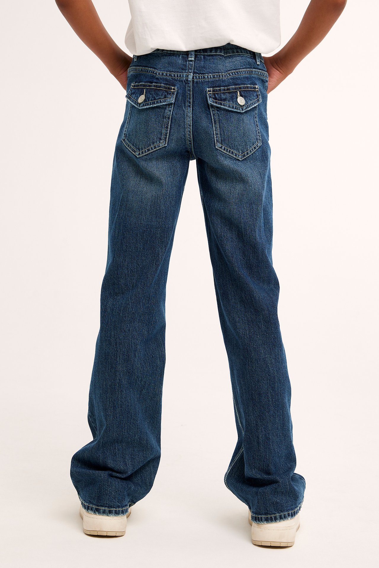 Widok z tyłu dziecka w ciemnych jeansach bootcut z klapami, białym T-shircie i jasnych sneakersach.