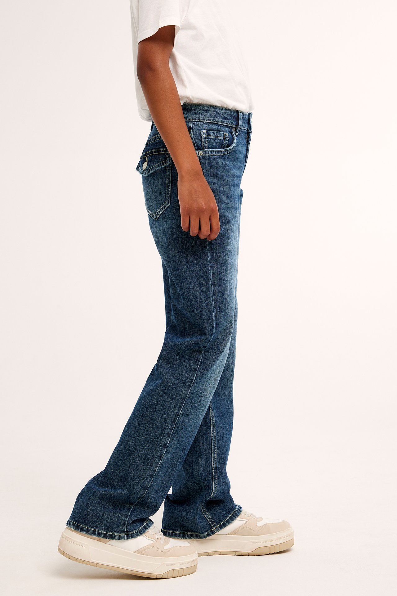 Dziecko z boku w ciemnych dżinsach bootcut z klapami, białym t-shircie i beżowych sneakersach.
