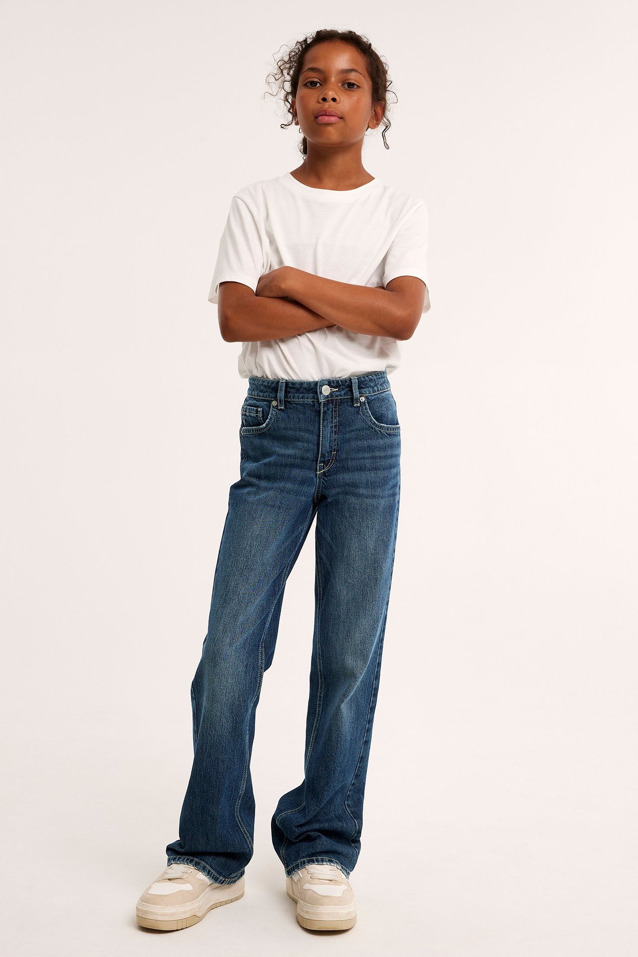 Hel frontvy av ett barn i mörkblå bootcut-jeans, en vit t-shirt och ljusbeige sneakers.