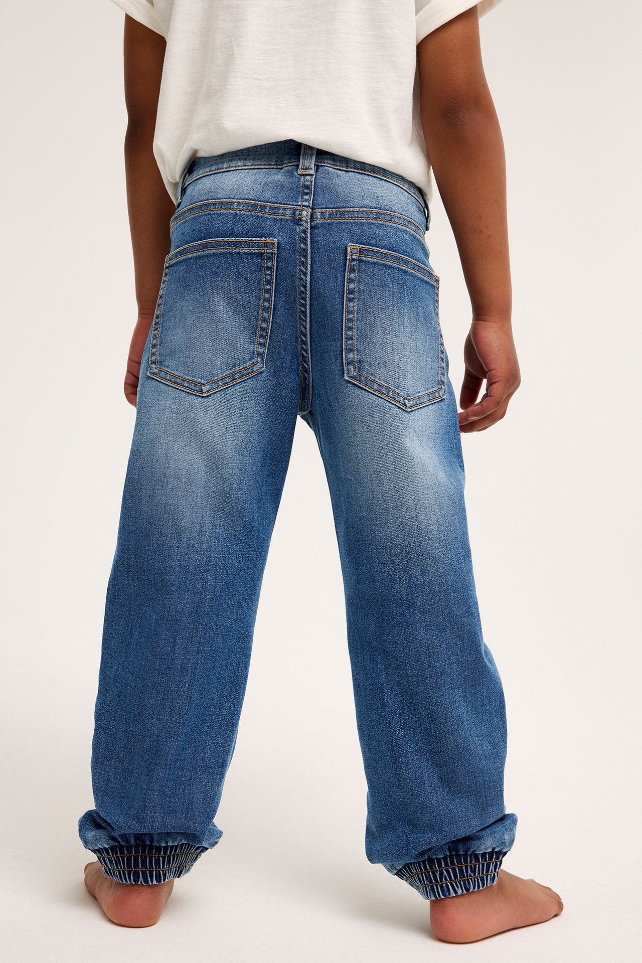 Bakifrån, ett barn i avslappnade blå denimjeans med resår i benslut och en vit t-shirt.