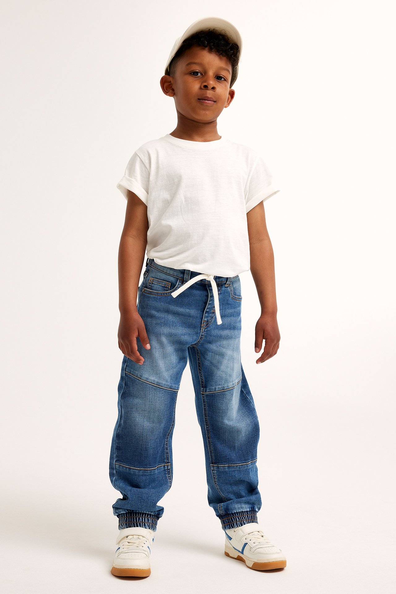 Framifrån: ett barn i blåa relaxed fit jeans med knäförstärkningar, vit t-shirt, beige keps och vita sneakers.