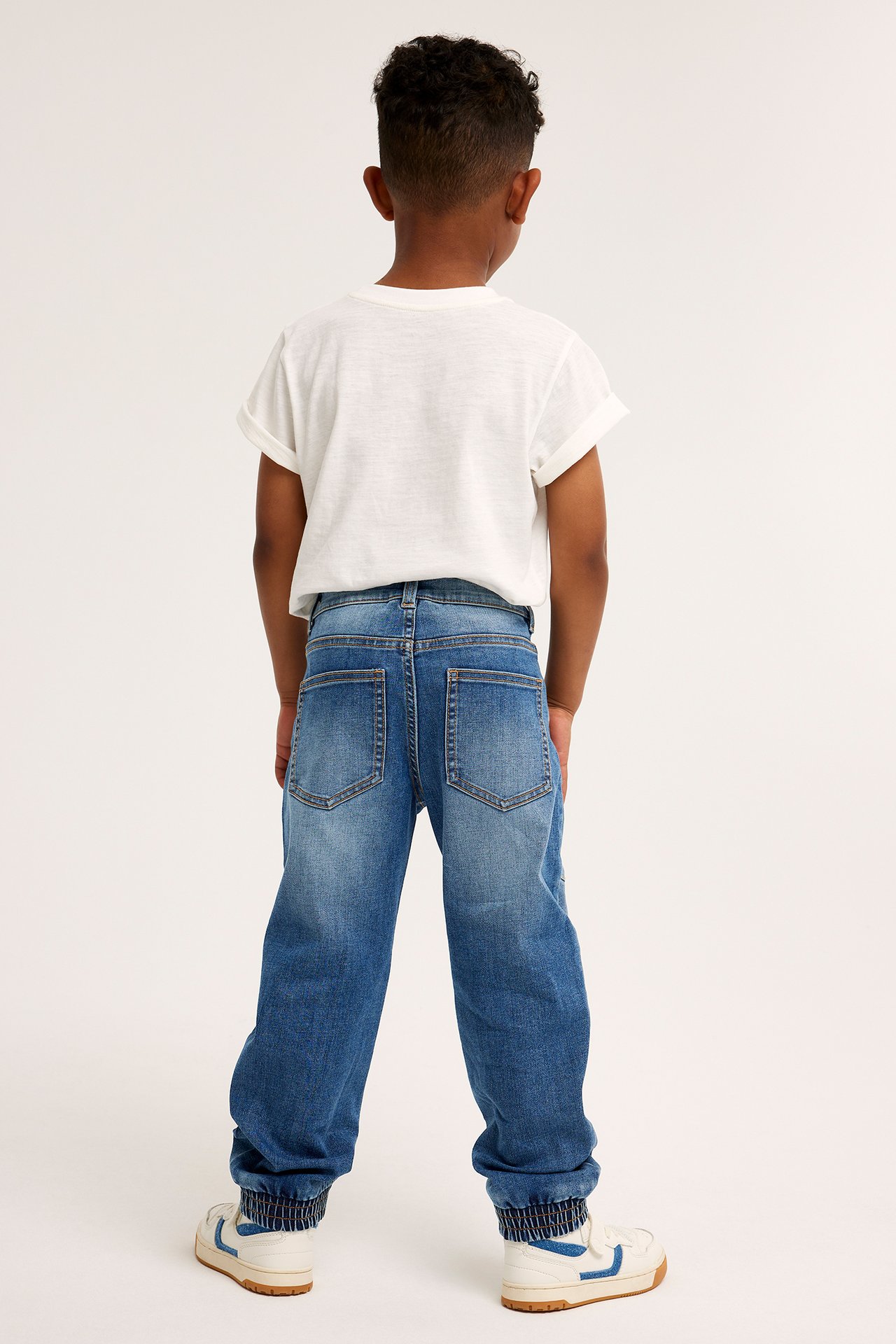 Bakifrån, ett barn i blåa relaxed denimjeans med resår i benslut, vit t-shirt och vita sneakers.