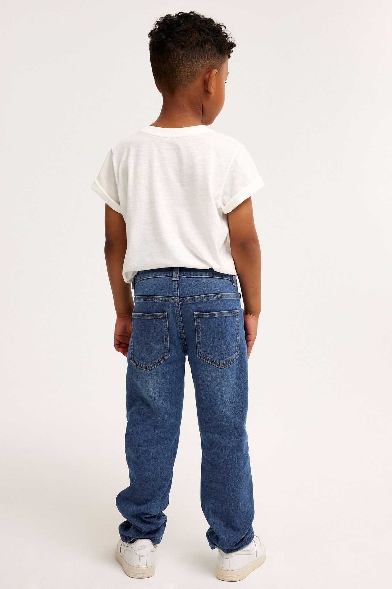 Bakfra, et barn i blå rette denimjeans, en hvit t-skjorte og hvite joggesko.