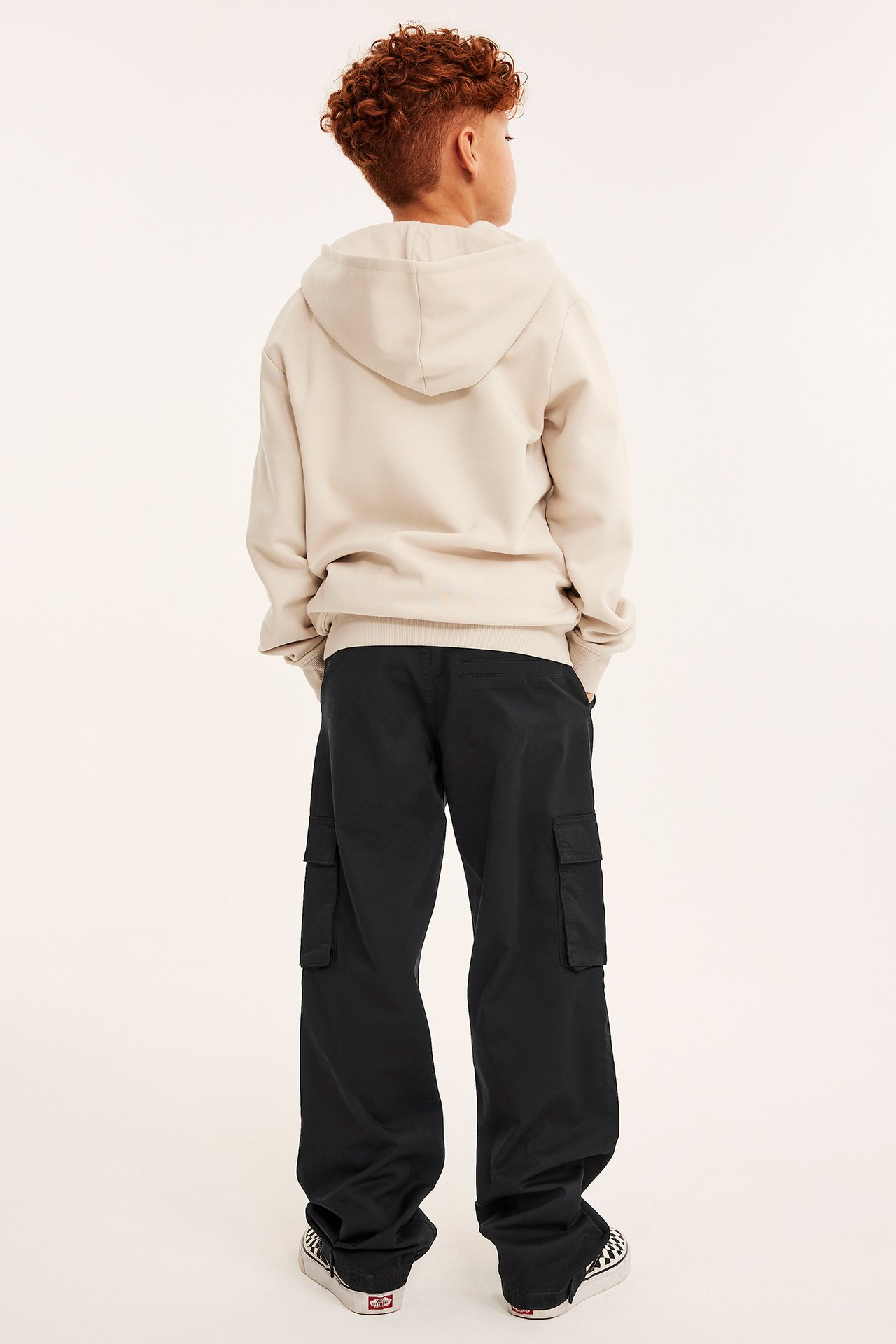Bakifrån på ett barn i svarta lösa cargobyxor med sidofickor, en ljusbeige huvtröja och rutiga sneakers.