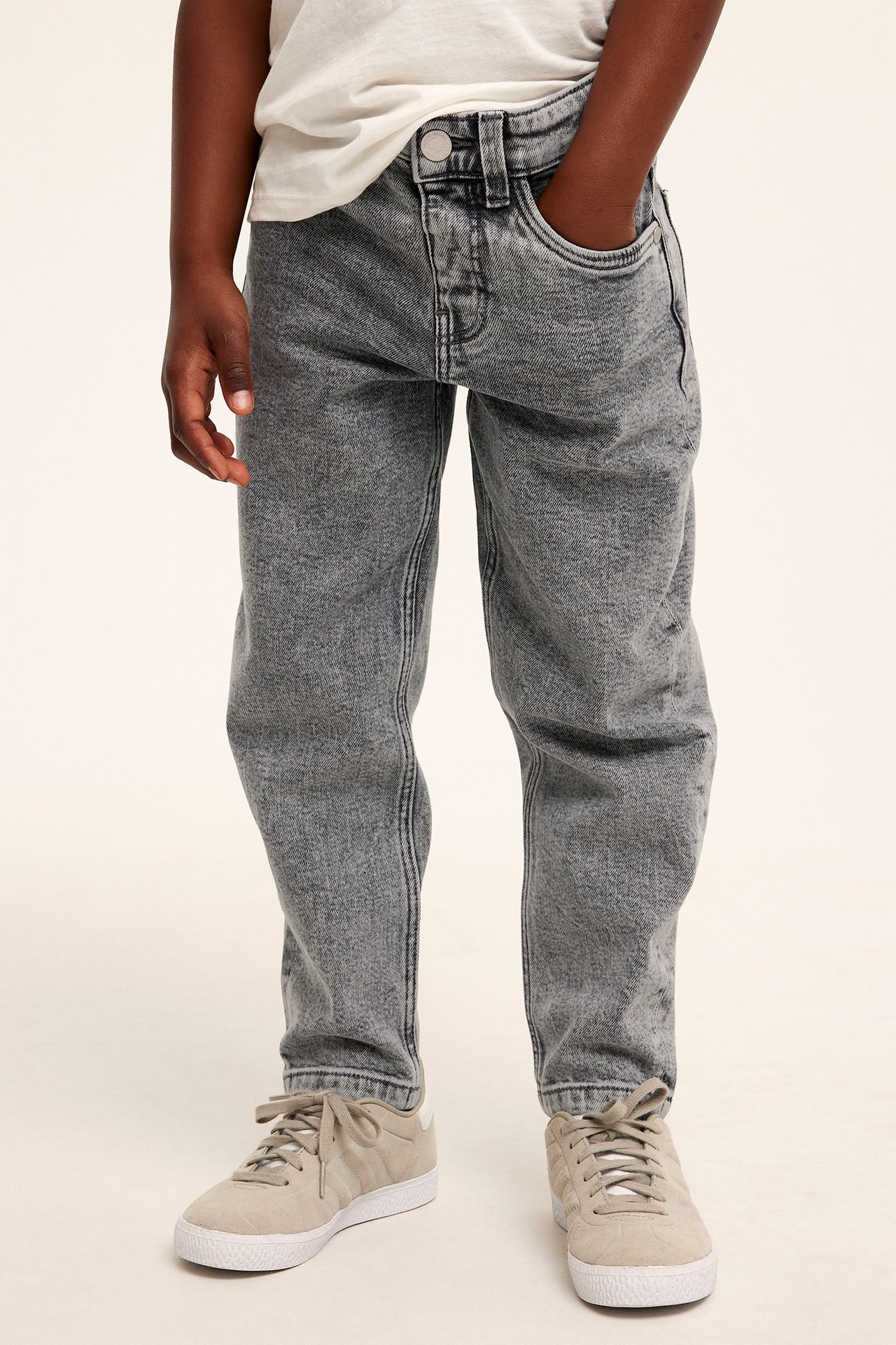 Dziecko w jasnych czarnych jeansach tapered z zatrzaskiem, białym T-shircie i beżowych sneakersach.