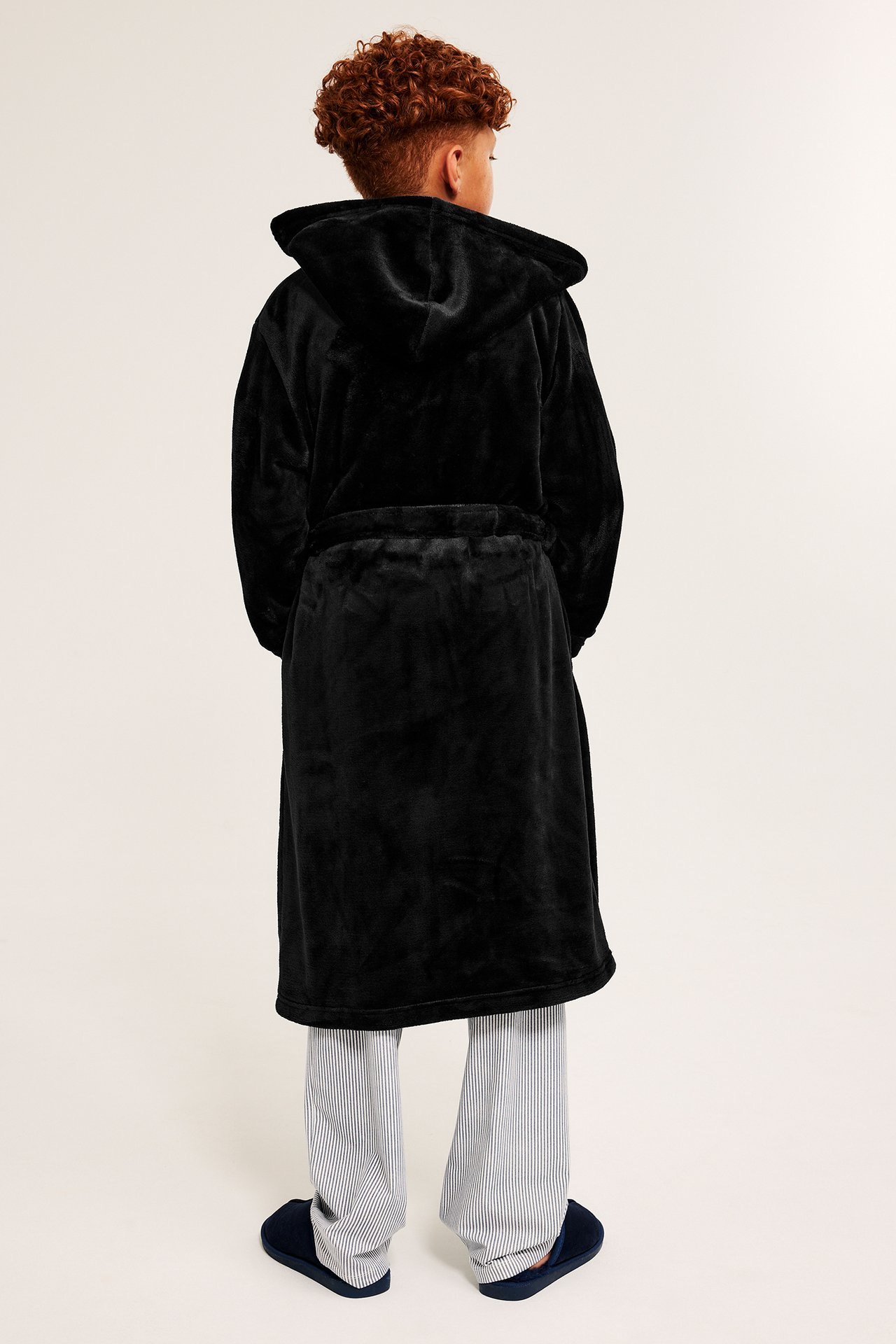 Bakifrån på ett barn i svart huvmorgonrock i fleece, över randiga pyjamasbyxor och mörkblå tofflor.