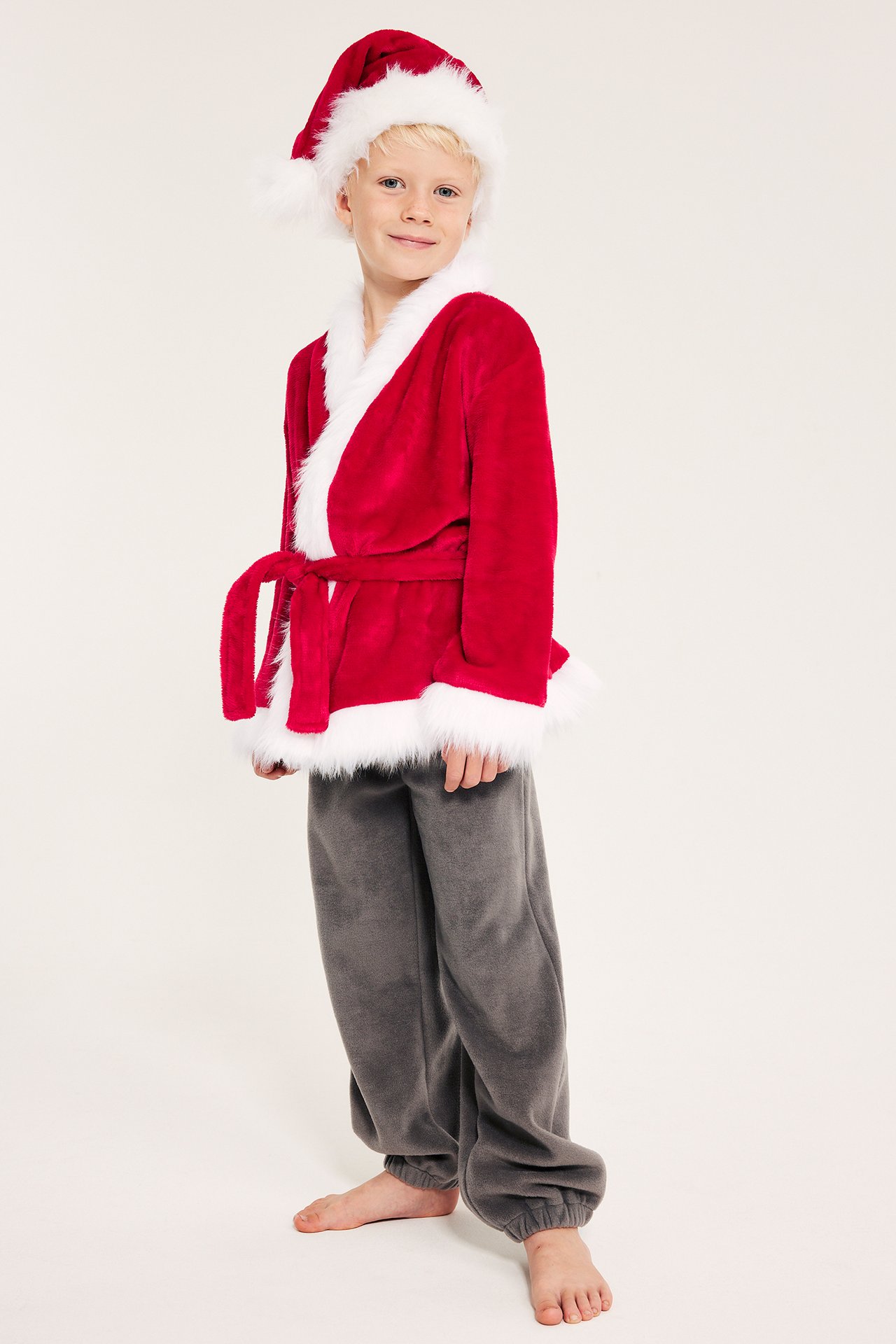 Barn i rødt fleece santa-pysjamas med hvite pelsdetaljer og grå bukser.