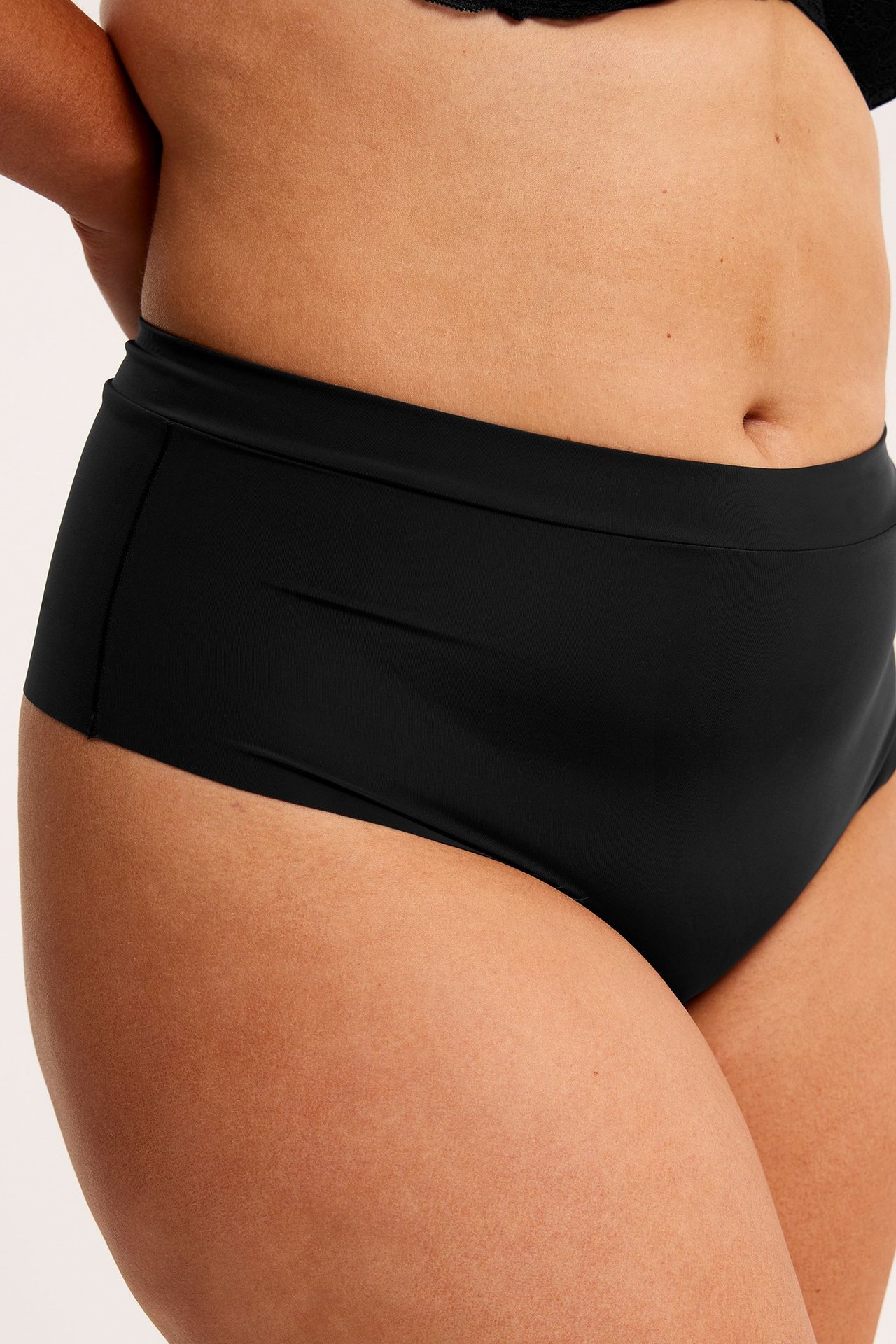 Lähikuva edestä naisesta, jolla on mustat korkeavyötäröiset mikrokangassongit ja mustat rintaliivit.