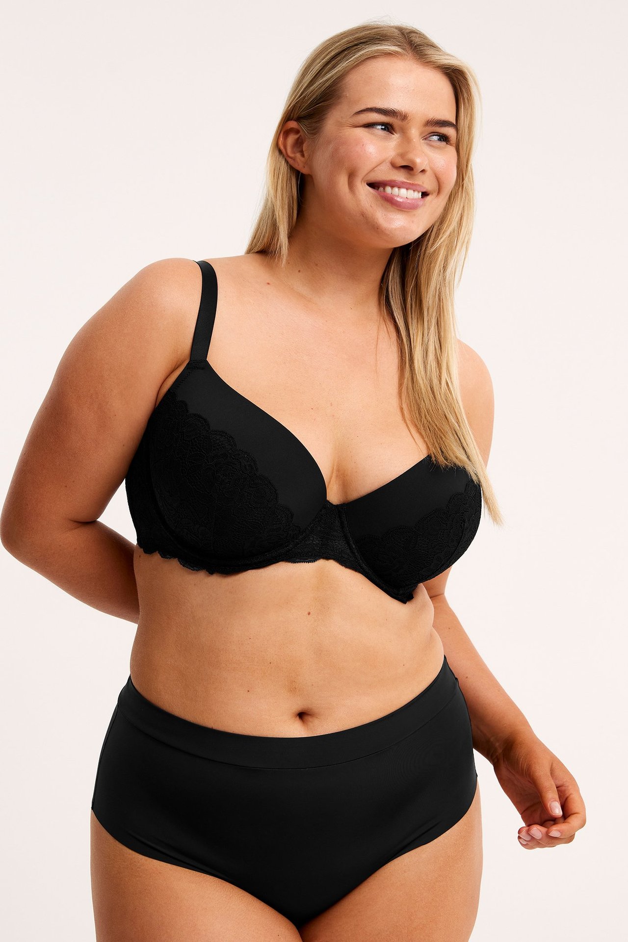 Frontbilde av en smilende kvinne i svart blonde-BH og glatte svarte høy-midjede truser.