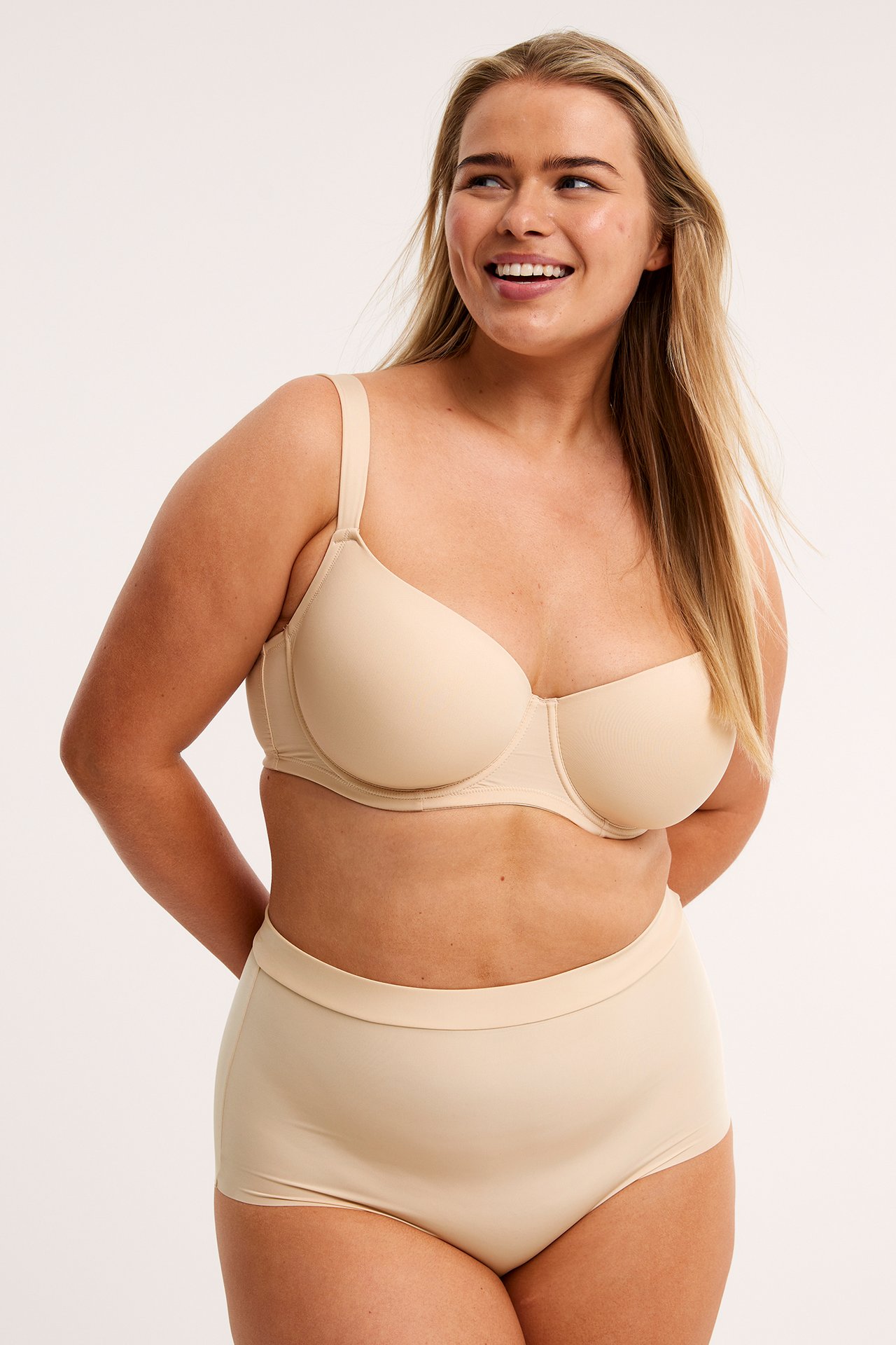 Frontbilde av smilende kvinne i beige sømløse hipstertruse med elastisk linning og matchende BH.