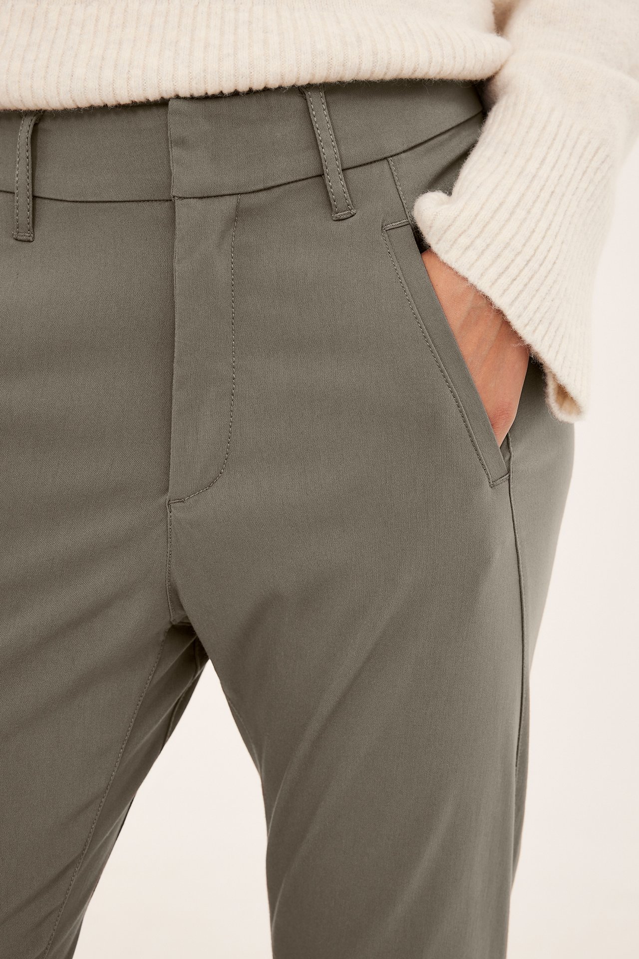 Nærbilde forfra av en kvinne i ensfargede khaki slim-fit bukser med en kremfarget ribbestrikket genser.