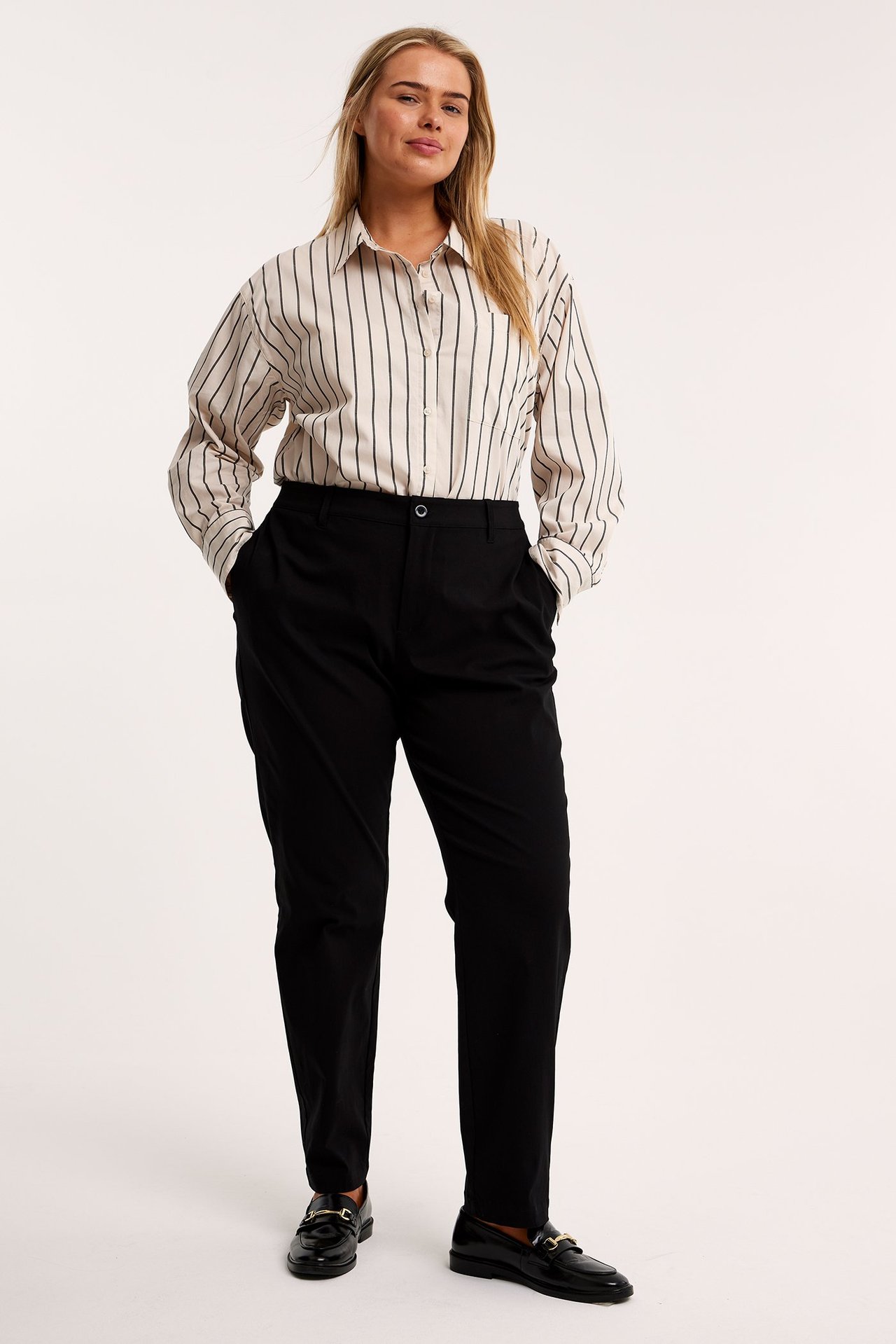 Widok z przodu: kobieta w czarnych spodniach garniturowych, beżowej koszuli w paski i czarnych mokasynach.