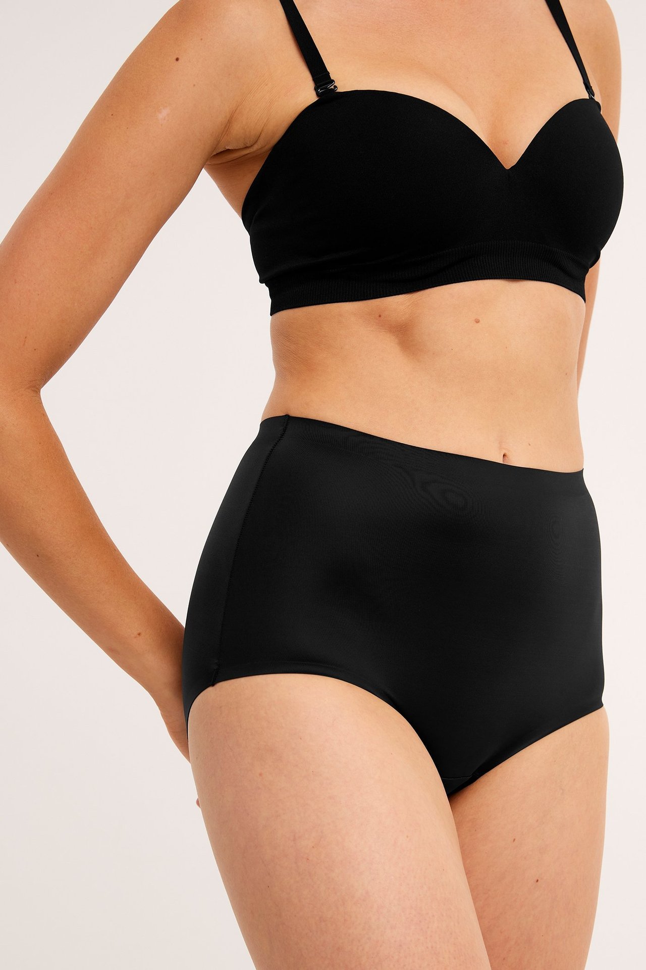 Sned framifrån-vy av en kvinna i högmidjade svarta shapingtrosor och en svart axelbandslös BH.