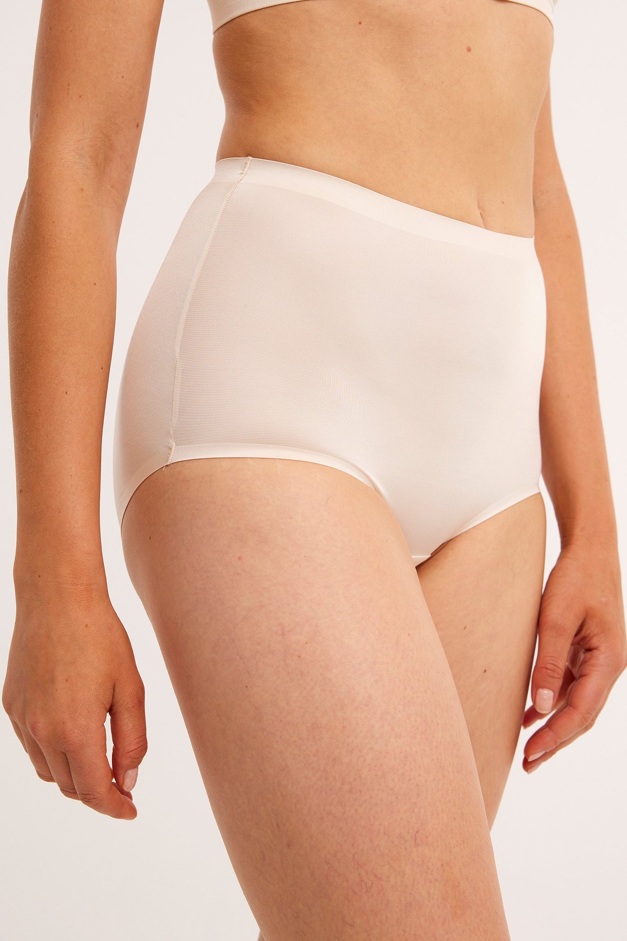 Sidebilde av en kvinne i høy midje beige shapingtruser i satinmicro og en matchende BH-topp.