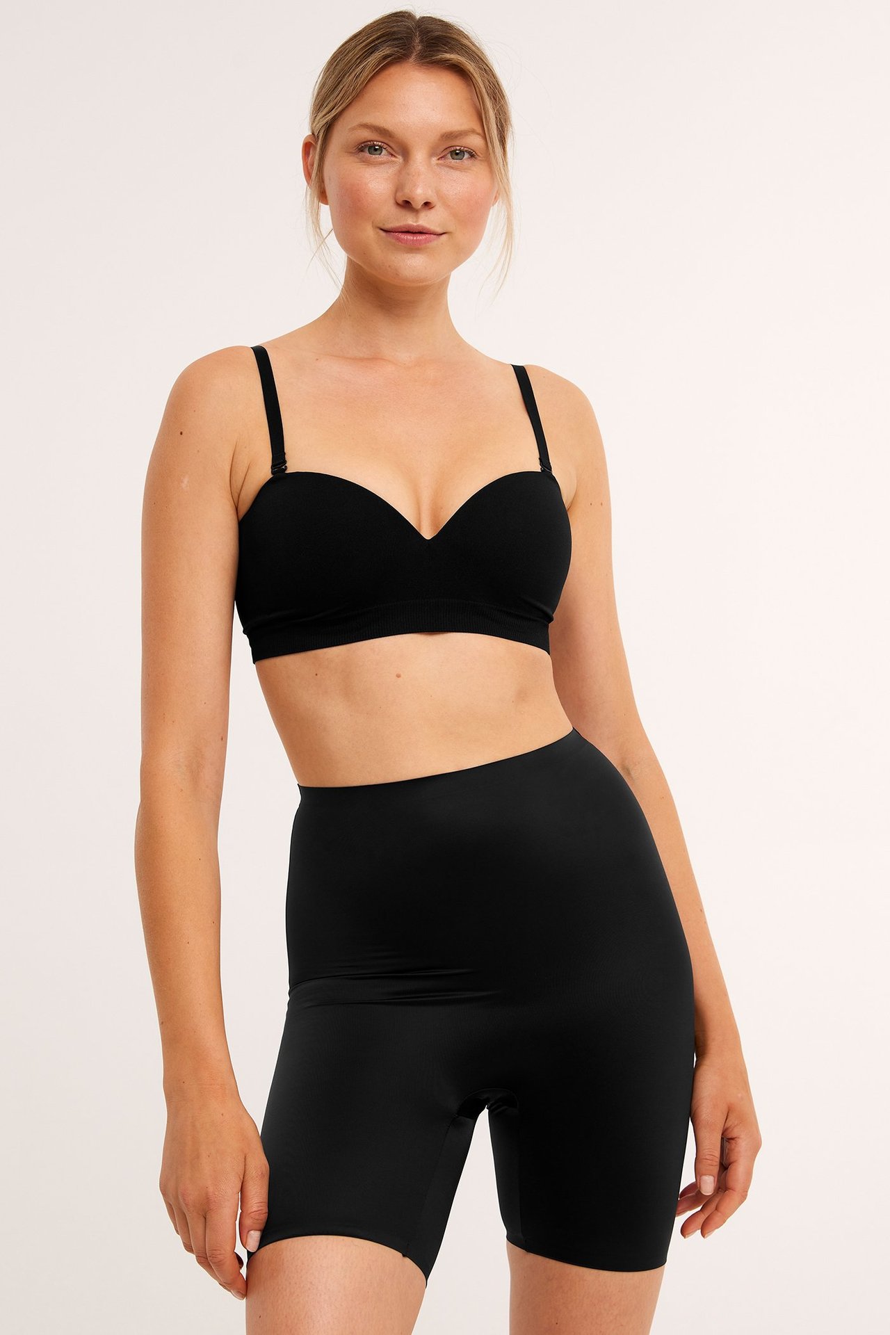 Frontbilde av kvinne i svarte høytlivs shaping-shorts i satinmicro og svart stroppeløs BH.