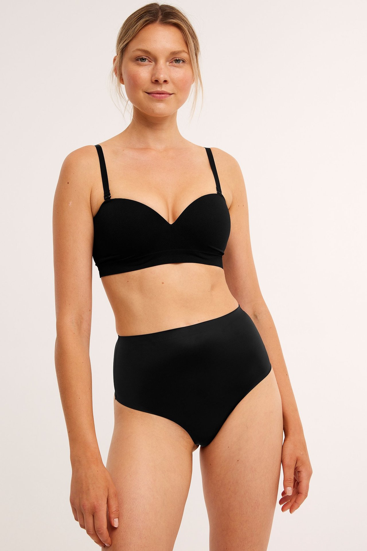 Forfra, kvinne i svart høytlivs shaping-stringtruse og svart stroppeløs BH.