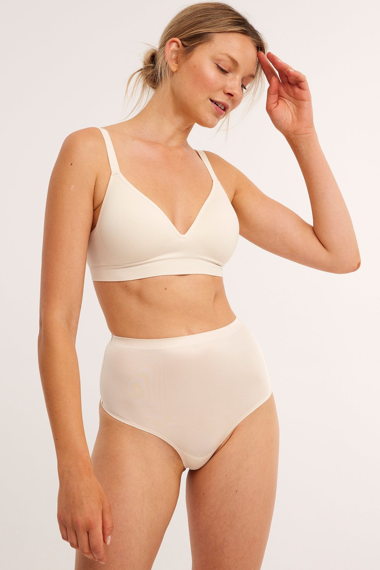 Framifrån, kvinna i högmidjad beige satinmicro stringtrosa och matchande mjuk-bh.