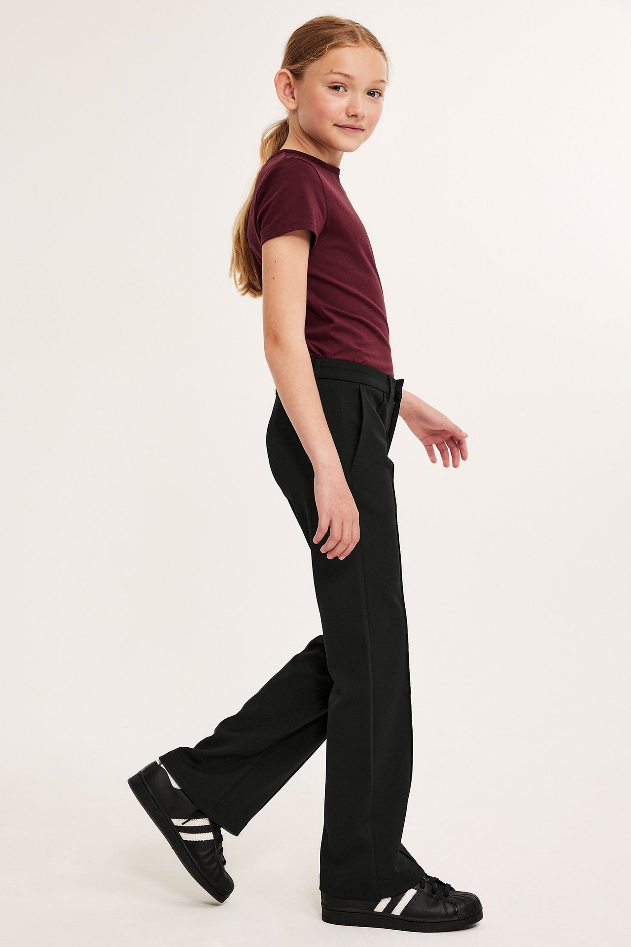 Widok z boku: Dziewczynka w czarnych dżersejowych spodniach flare z przeszyciem, bordowym t-shircie i czarnych sneakersach.