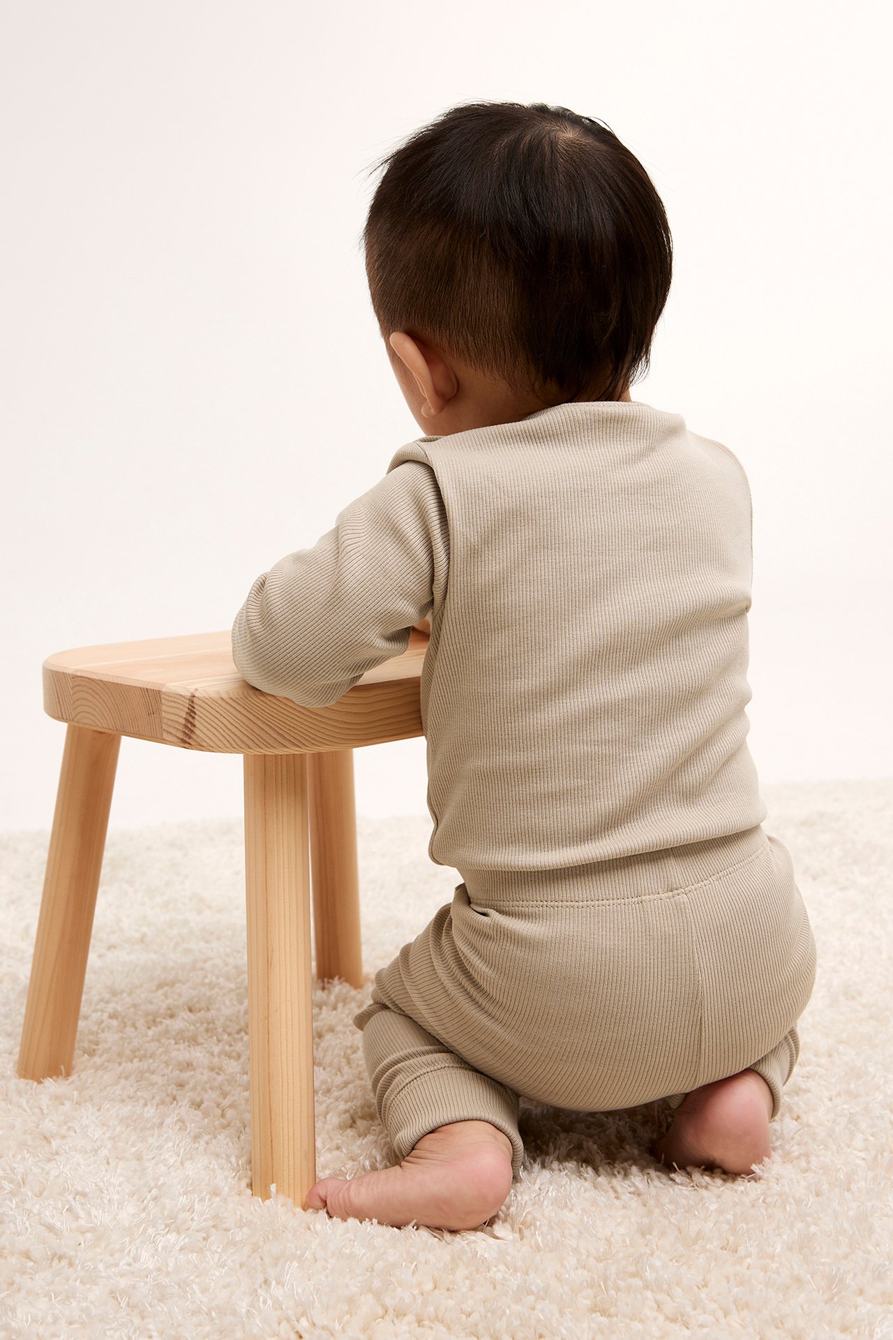 Vauva takaa, yllään beige ribattu pitkähihainen body, polvillaan puisen jakkaran vieressä.