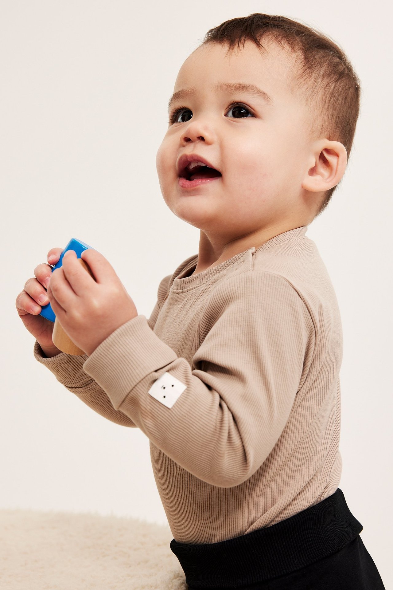 Sivukuva vauvasta beigessä ribatussa pitkähihaisessa bodyassa olkapään napeilla, pitäen lelua.