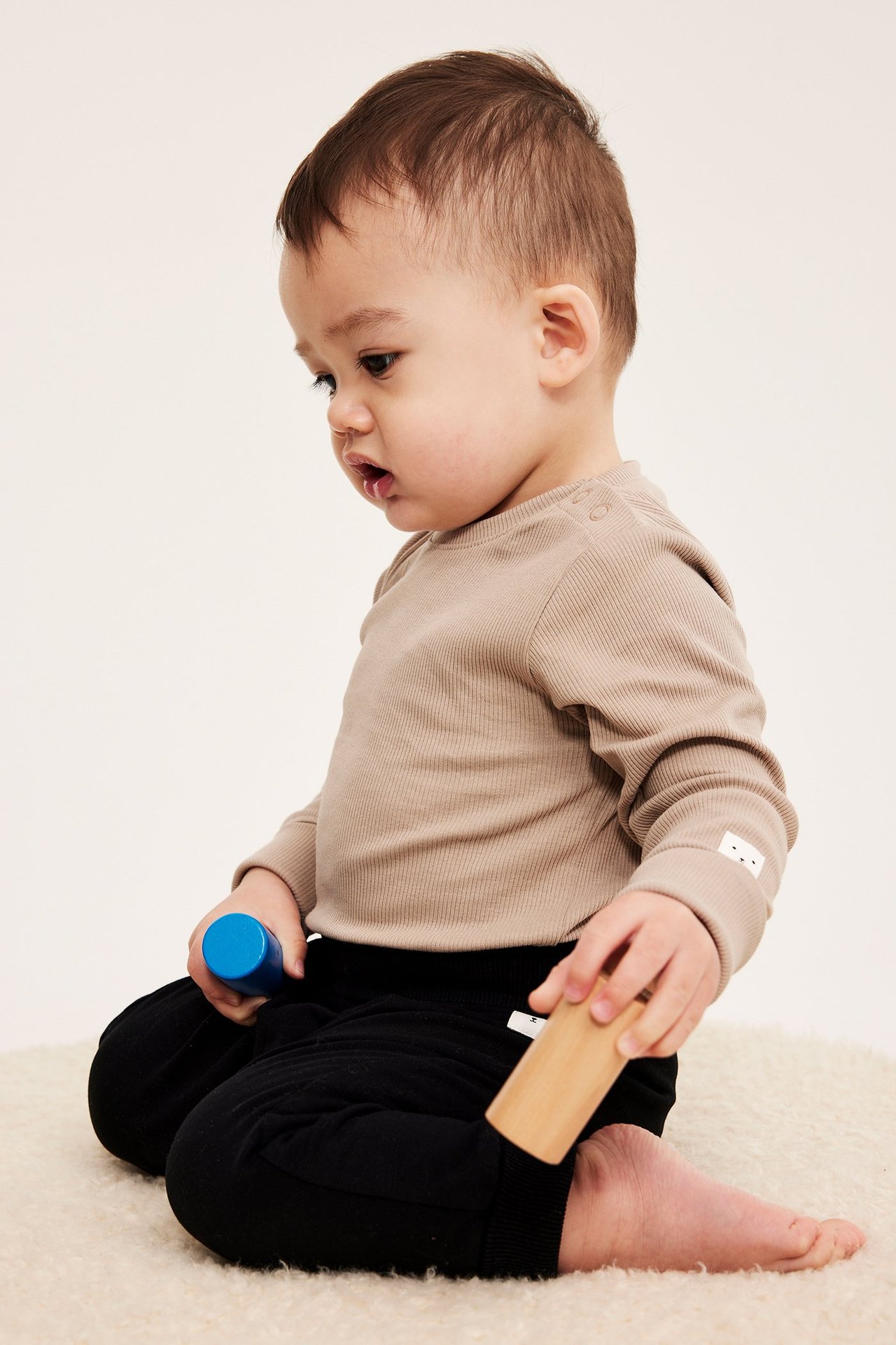 Sivukuva vauvasta beigessä ribatussa pitkähihaisessa bodyssä ja mustissa housuissa, pitäen leluja.