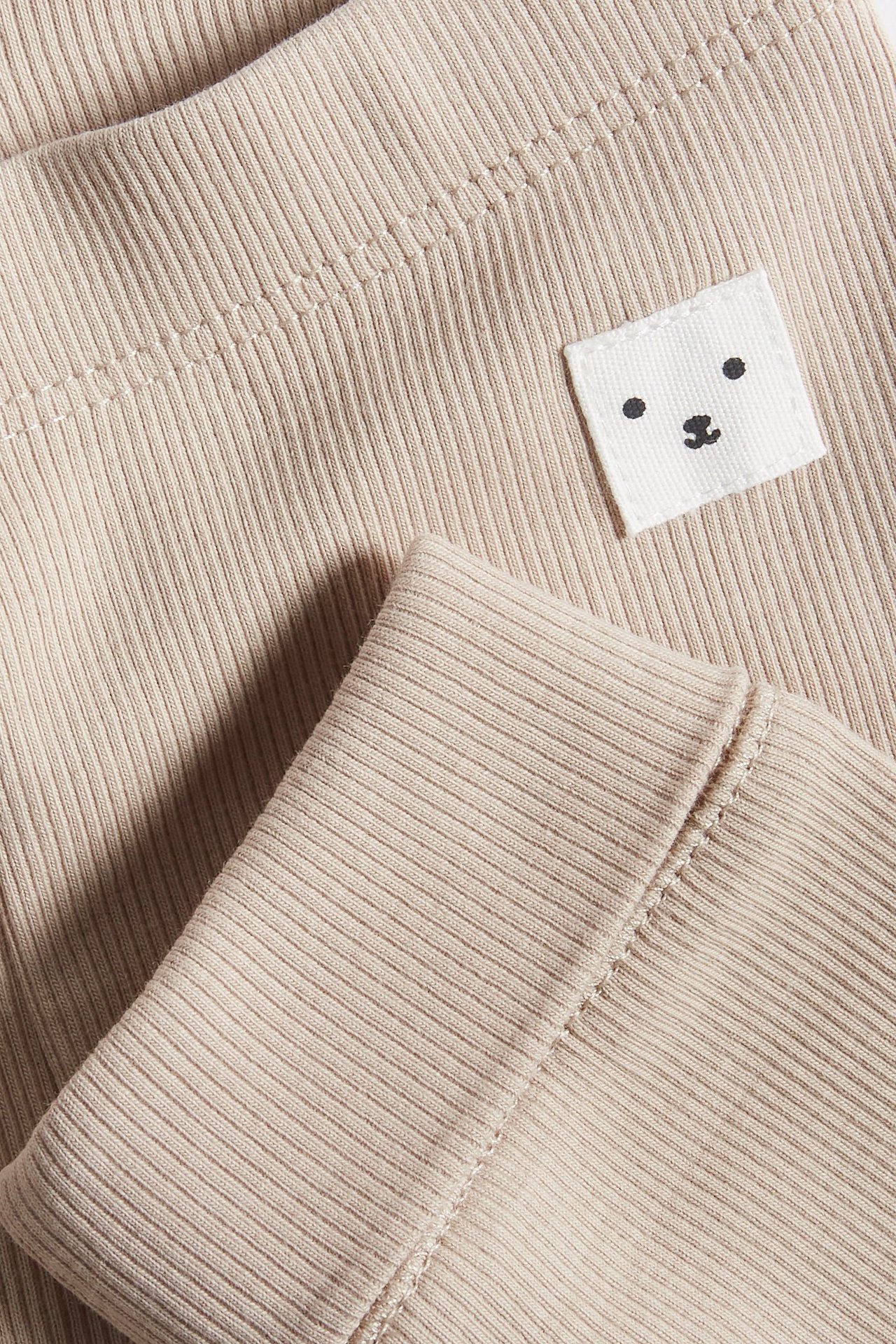 Närbild av beiga ribbade baby leggings med en vit björnansiktslapp och generösa muddar.