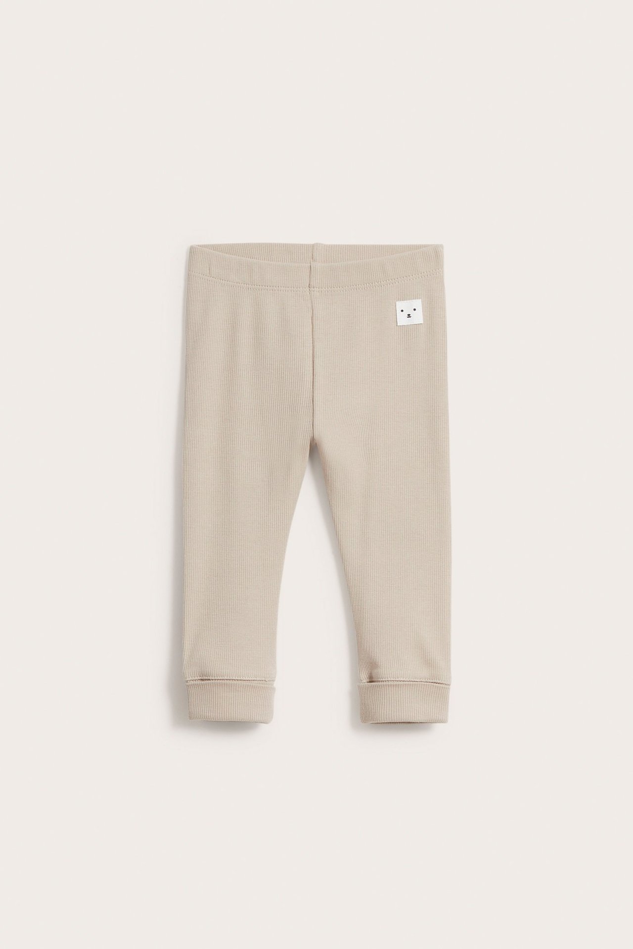 Frontvy av beiga ribbade baby leggings med resår i midjan, vikta muddar och en liten björnetikett.