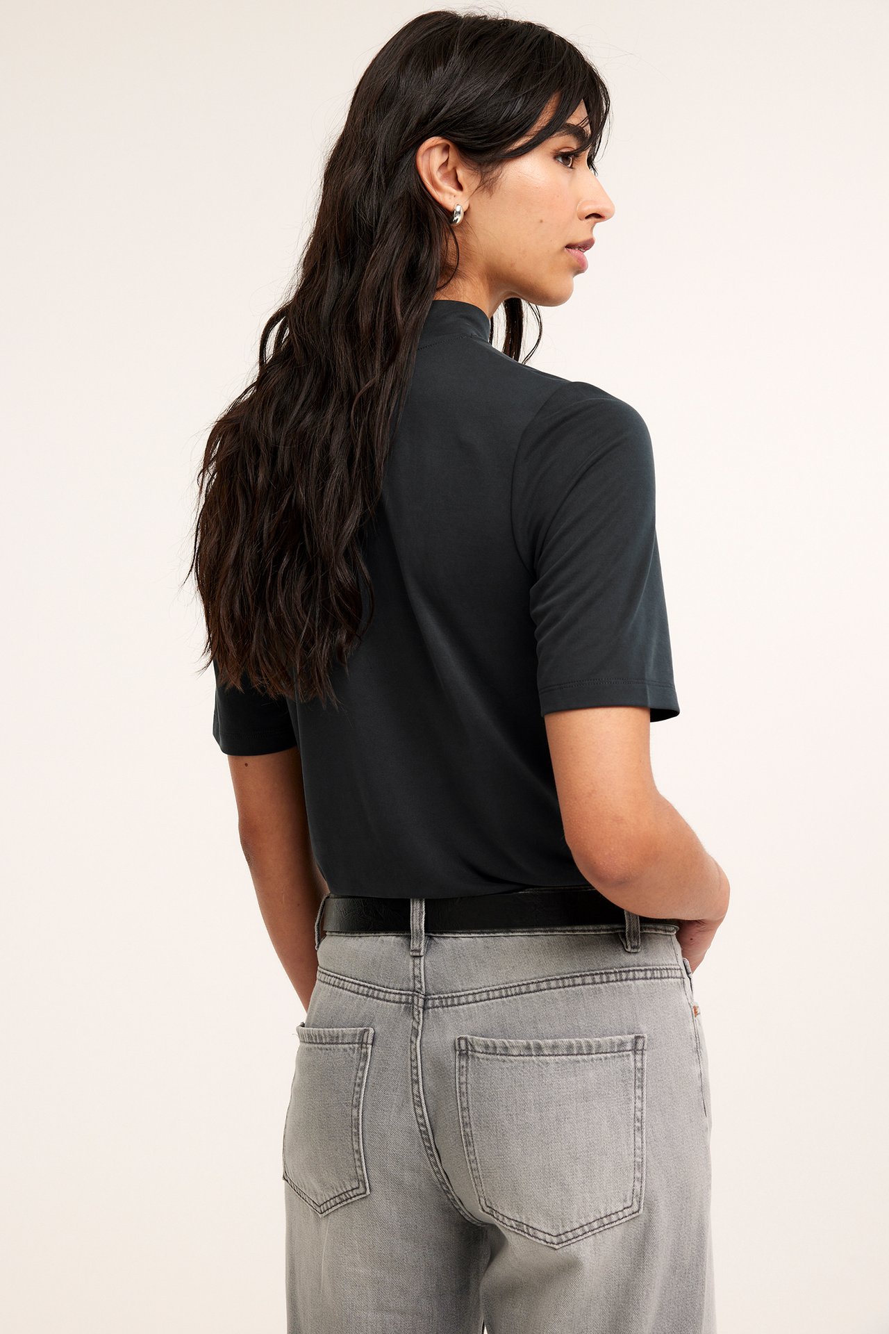 Sidovy av en kvinna i svart kortärmad modalpolotopp, grå jeans, svart bälte och silverörhängen.