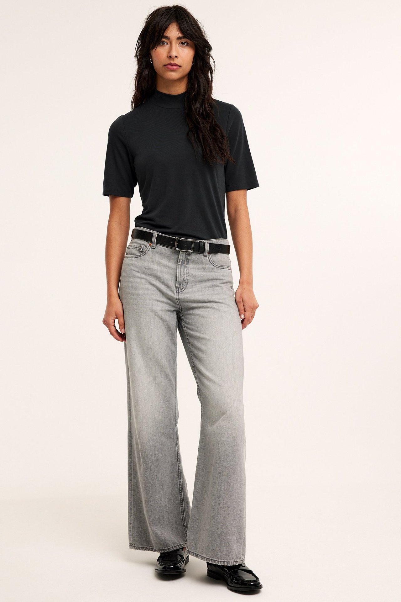 Frontvisning: kvinne i svart kortermet modal turtleneck t-skjorte, grå vide jeans, svart belte og loafers.