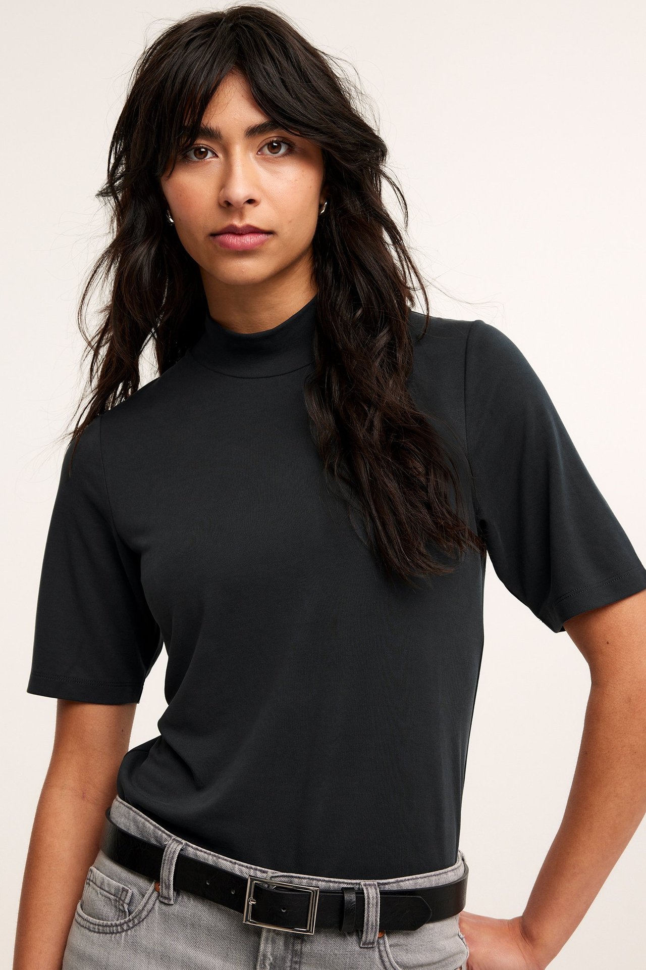 Frontbilde: kvinne i svart modal kortermet polotopp, lysegrå jeans og svart belte.