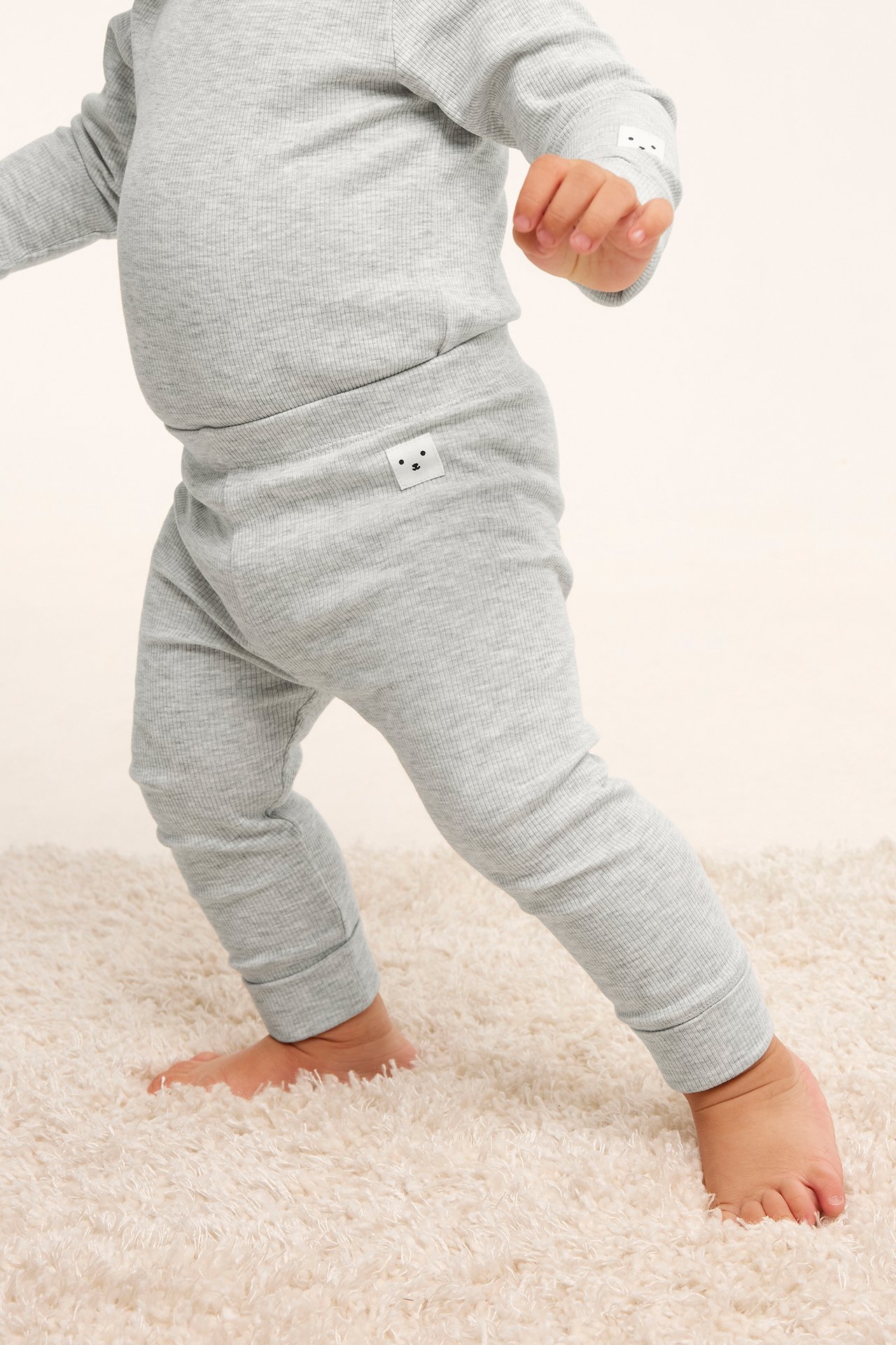 Närbild på baby i ljusgrå ribbade leggings och matchande topp, stående på en lurvig matta.
