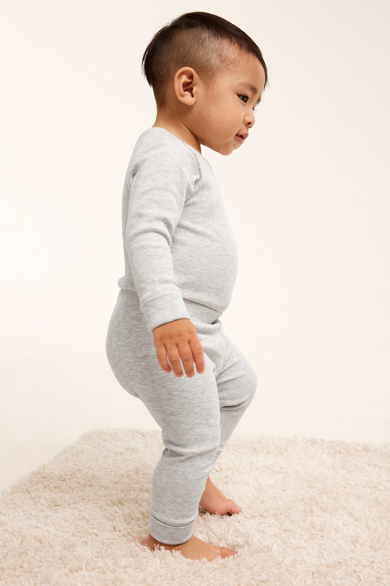 Sidovy av en baby i ljusgrå ribbade leggings och matchande långärmad topp på en matta.
