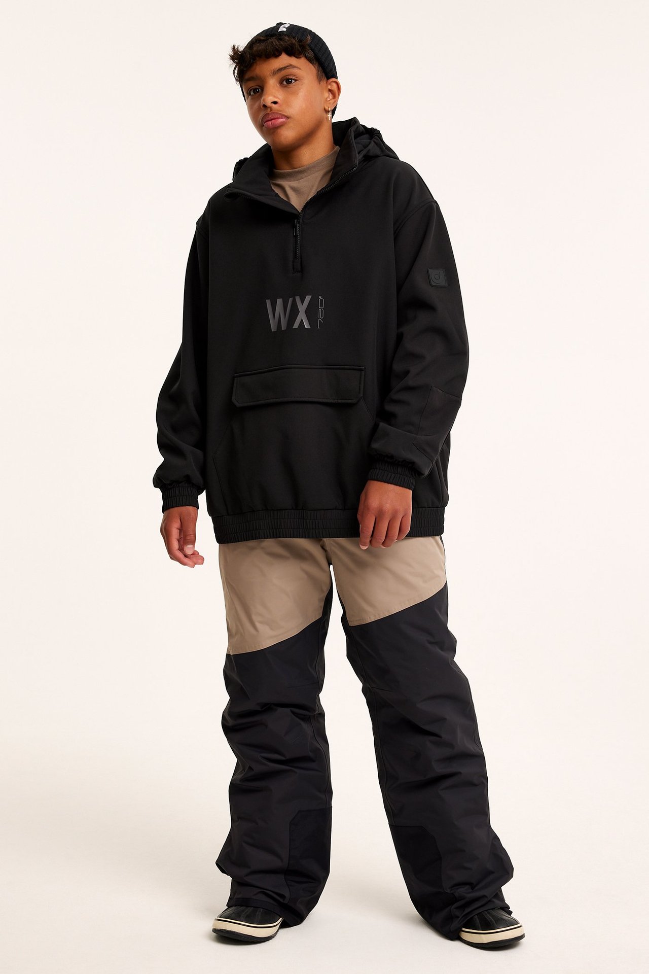 Widok z przodu: Dziecko w czarnej kurtce softshell, beżowo-czarnych spodniach narciarskich i czarnej czapce.