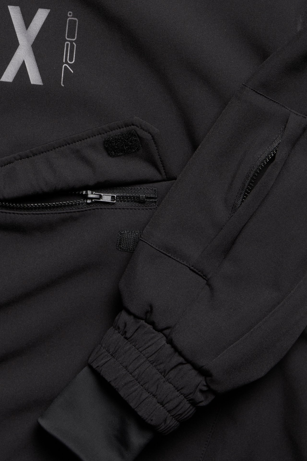 Lähikuva mustasta softshell-anorakista, jossa vetoketjullinen kengurutasku, joustava hihansuu ja heijastava painatus.