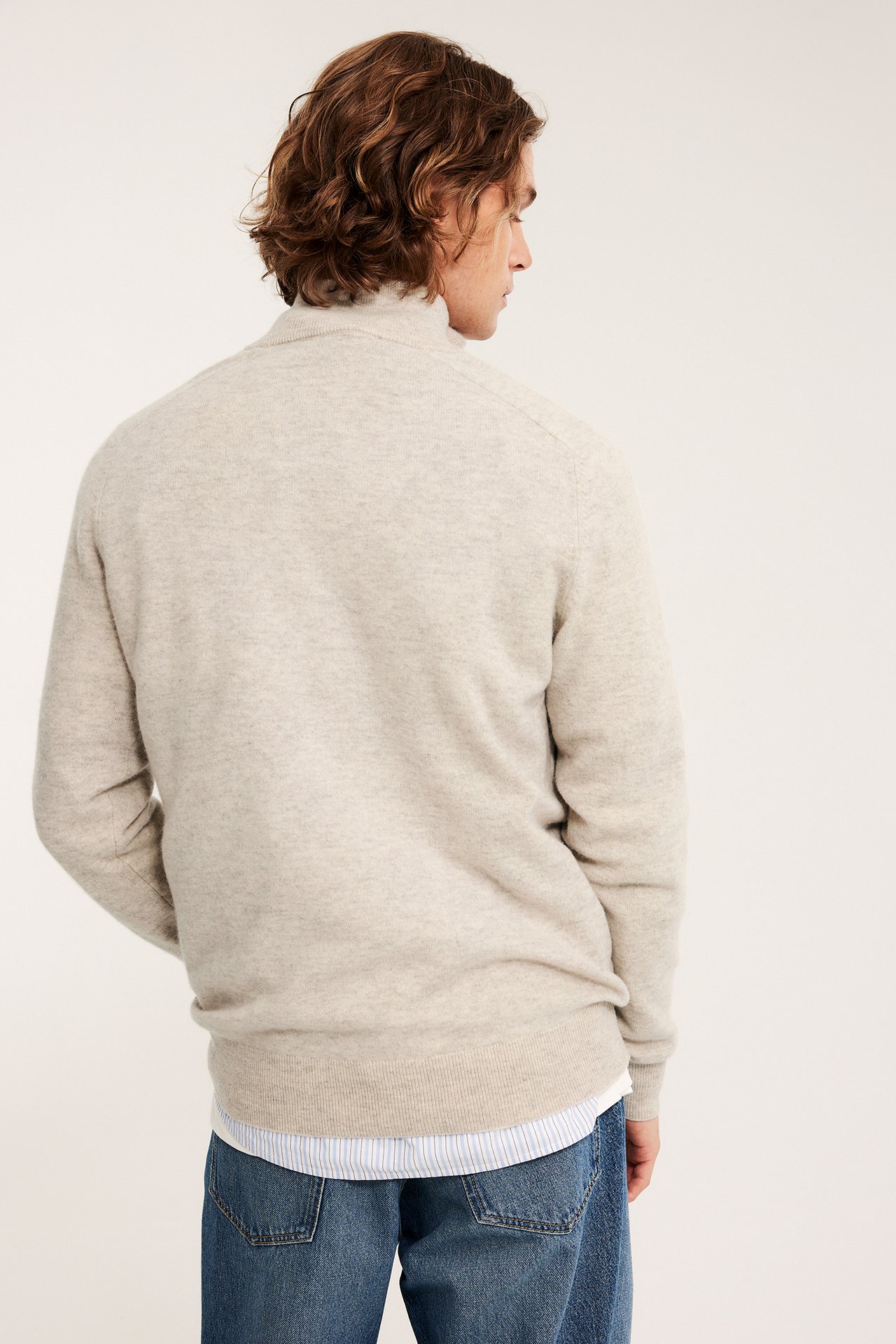 Miehen takakuva beige melange puolivetoketjullisessa villapaidassa raidallisen paidan ja farkkujen päällä.