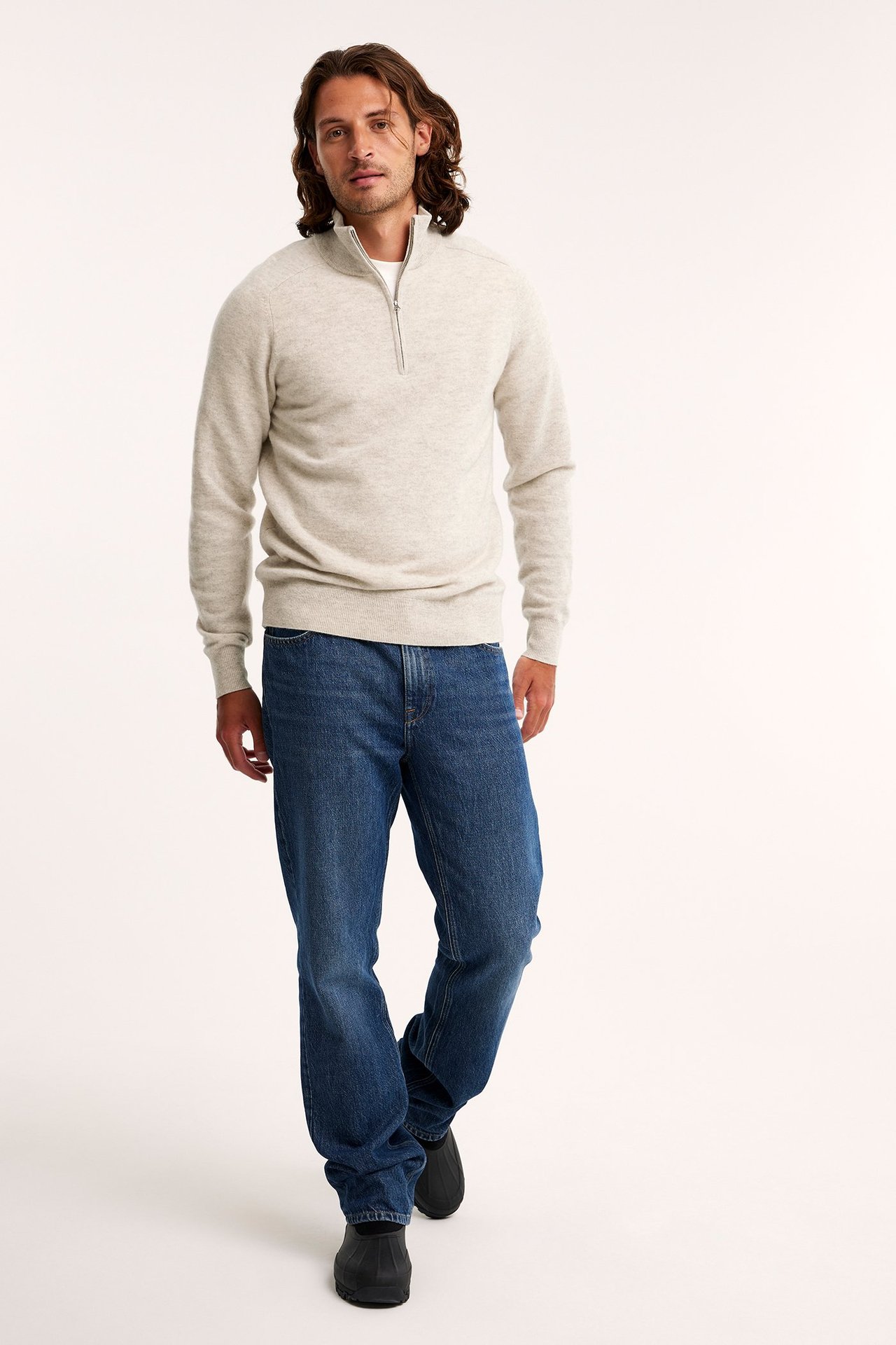 Frontvy av en man i beige melerad half-zip ulltröja, blå jeans och svarta skor.