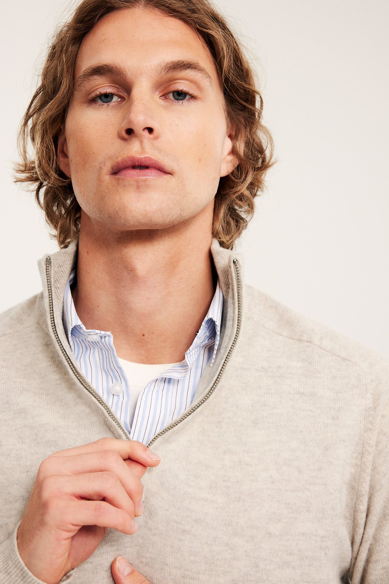 Zbliżenie: mężczyzna zapina beżowy melanżowy wełniany sweter z krótkim zamkiem, noszony na koszuli w paski.