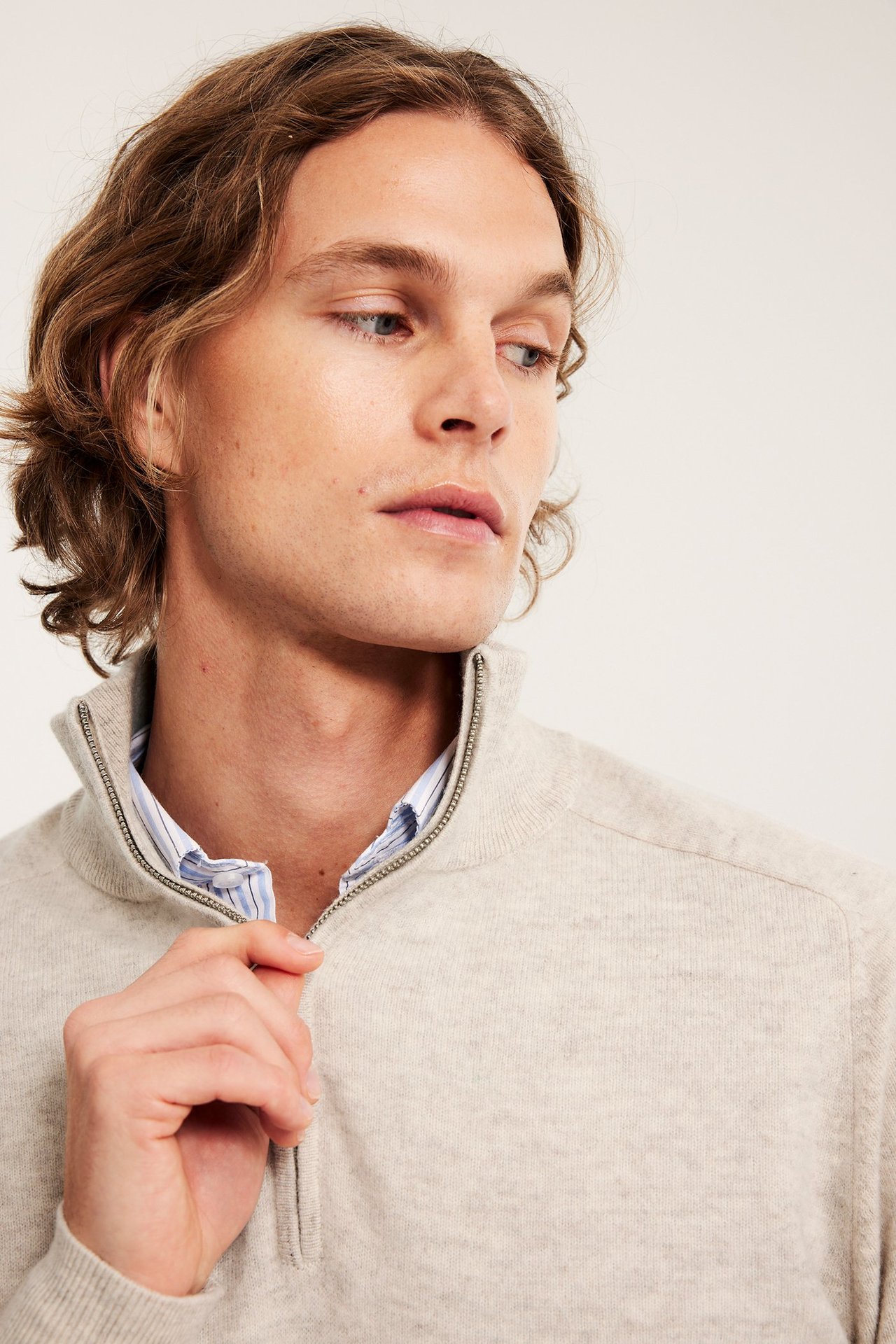 Zbliżenie mężczyzny w beżowym melanżowym wełnianym swetrze z półzamkiem nałożonym na jasnoniebieską koszulę w paski.